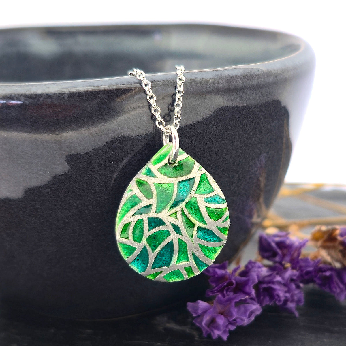 A handmade teardrop-shaped pendant necklace featuring a green enamel mosaic pattern with a silver chain, shown draped on a black bowl with dried purple flowers. Mijoux Jewellery.