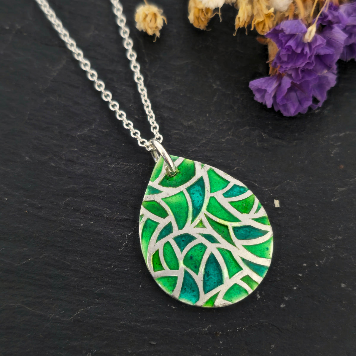 A handmade silver teardrop pendant necklace with a vibrant green enamel and silver mosaic leaf pattern, resting on a dark slate surface with dried flowers in the background. Mijoux Jewellery.