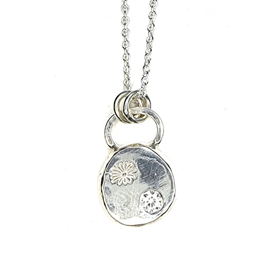 A round silver pendant with a daisy flower embossed on it and set with a clear CZ gemstone. On a silver chain.