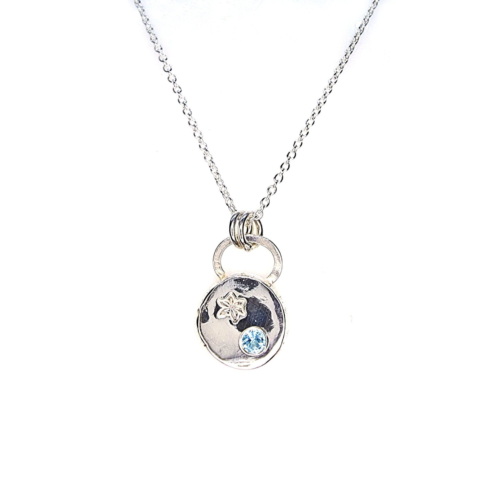 A silver round flat pebble pendant set with a round blue topaz gemstone (December birthstone) and engraved with a poinsettia flower (December birth flower) on a silver chain.