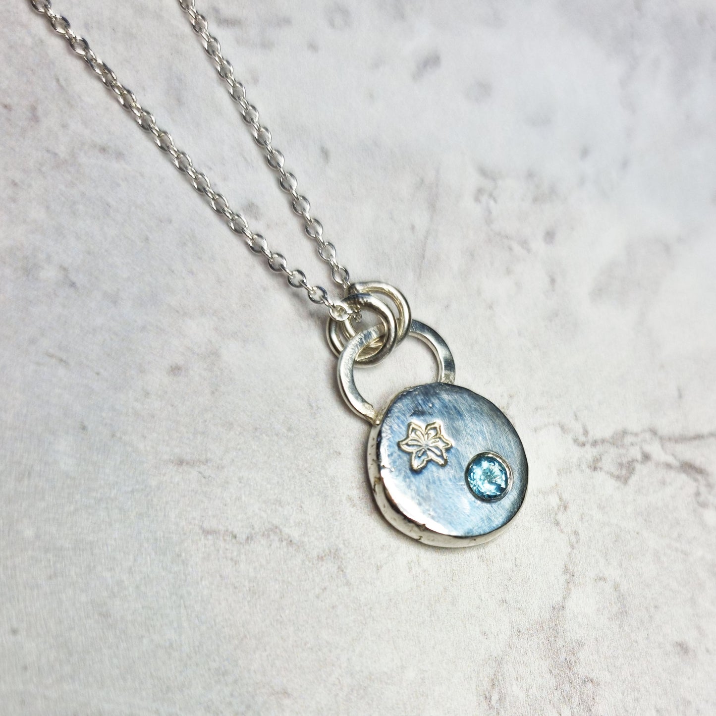 A silver round flat pebble pendant set with a round blue topaz gemstone (December birthstone) and engraved with a poinsettia flower (December birth flower) on a silver chain.