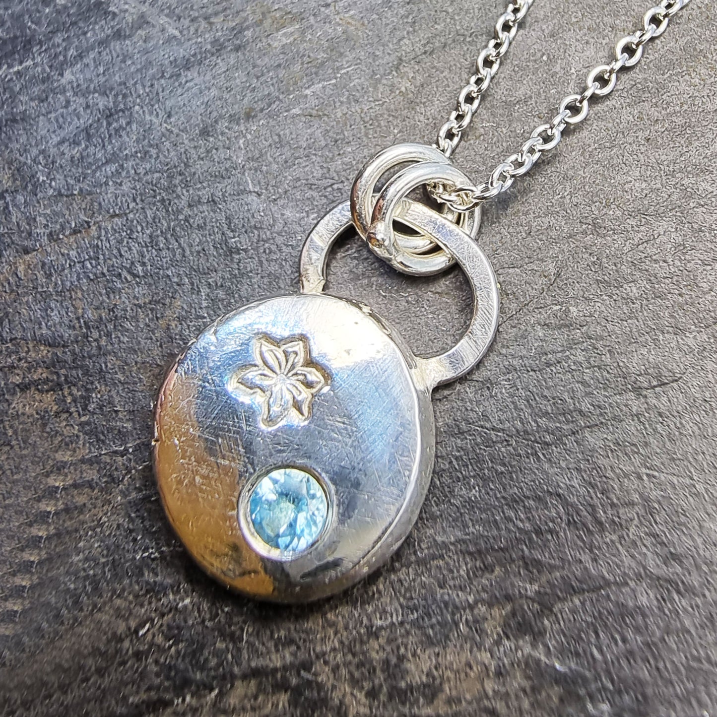 A silver round flat pebble pendant set with a round blue topaz gemstone (December birthstone) and engraved with a poinsettia flower (December birth flower) on a silver chain.