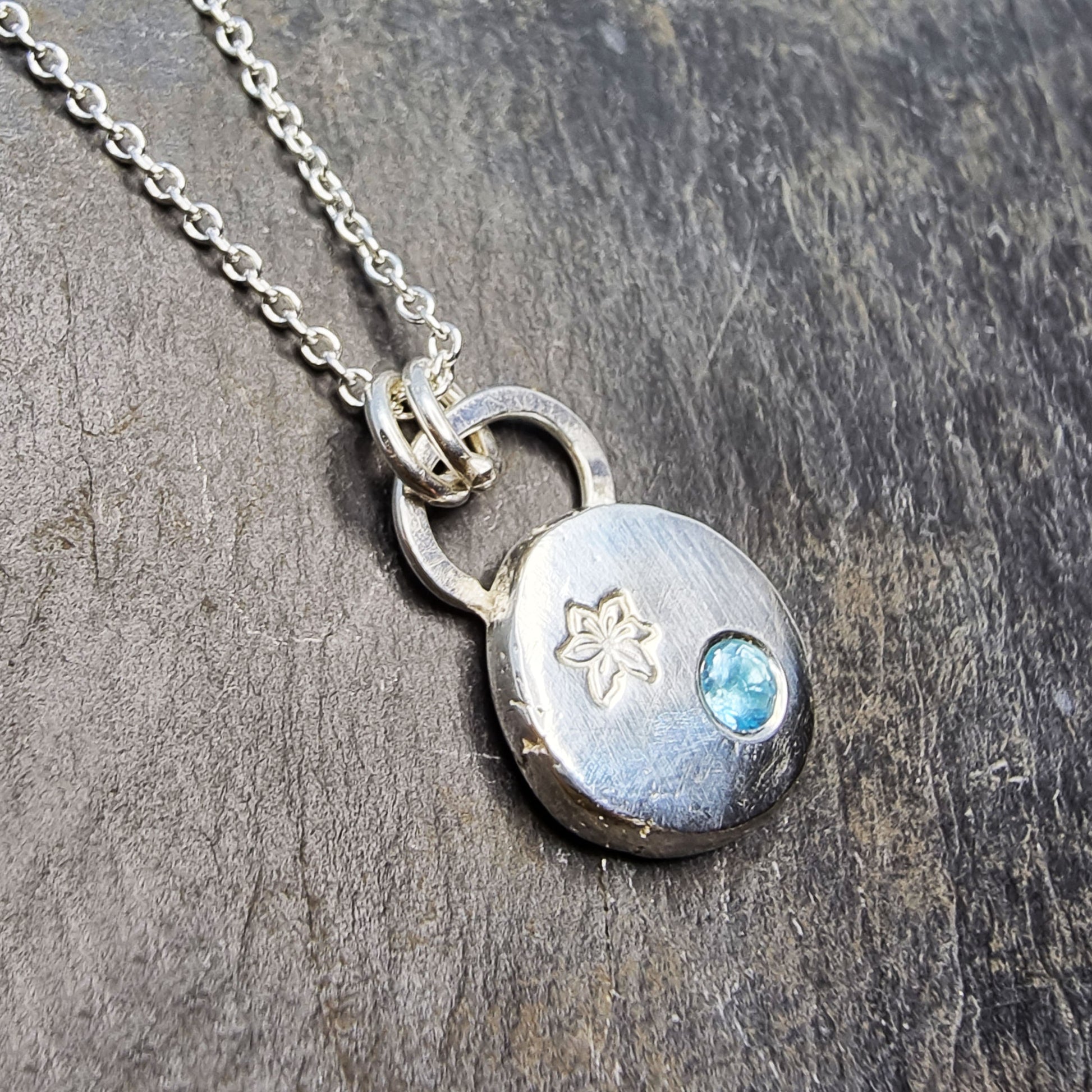 A silver round flat pebble pendant set with a round blue topaz gemstone (December birthstone) and engraved with a poinsettia flower (December birth flower) on a silver chain.