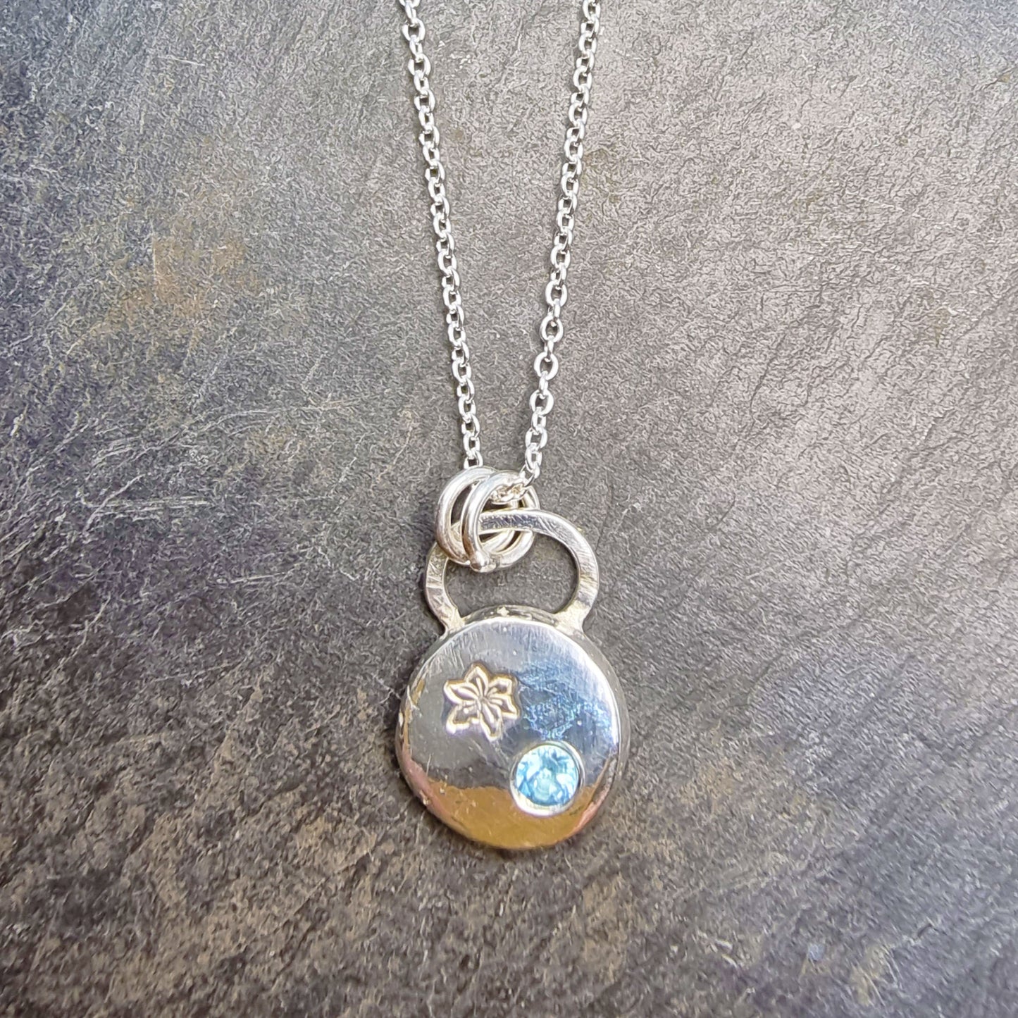 A silver round flat pebble pendant set with a round blue topaz gemstone (December birthstone) and engraved with a poinsettia flower (December birth flower) on a silver chain.