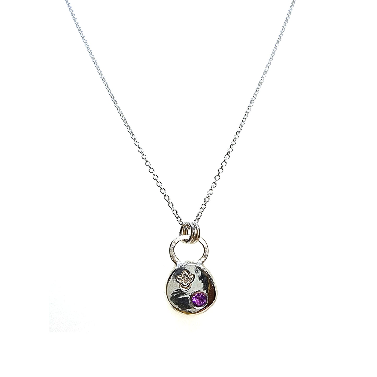 A round silver pendant with a violet flower (February birth flower) engraved on it and a round purple amethyst (February birthstone) flush set into it. On a silver chain.