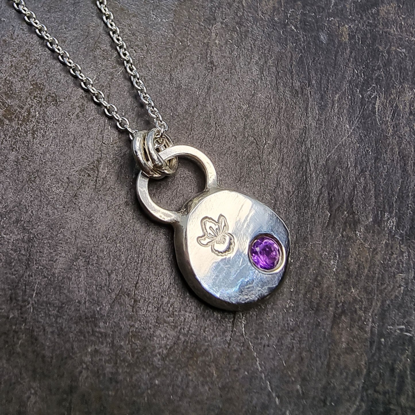 A round silver pendant with a violet flower (February birth flower) engraved on it and a round purple amethyst (February birthstone) flush set into it. On a silver chain.