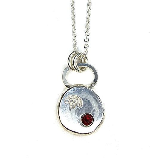 A round silver flat pebble pendant with a carnation flower engraved on it and a red garnet gemstone flush set into it. On a silver chain.