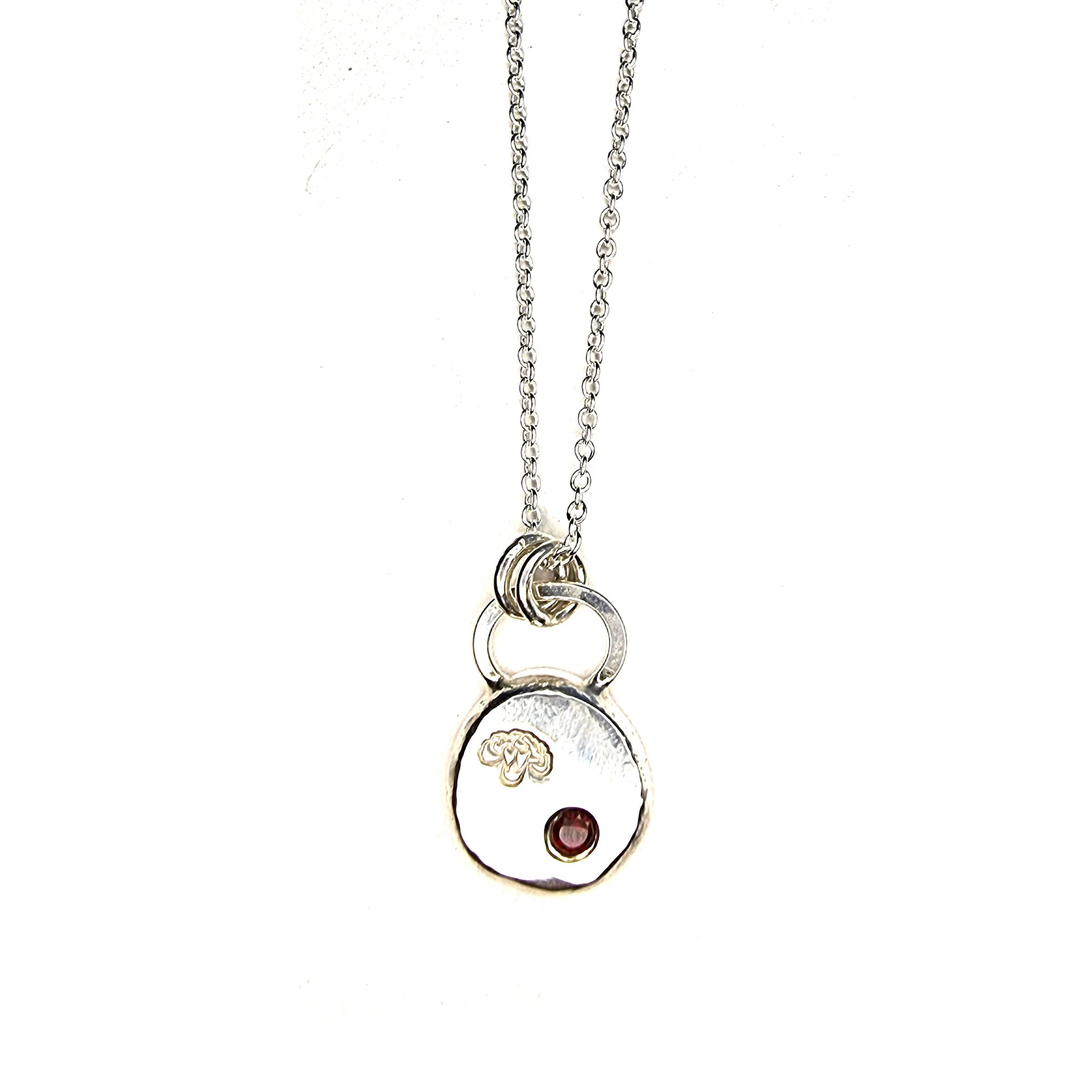 A round silver flat pebble pendant with a carnation flower engraved on it and a red garnet gemstone flush set into it. On a silver chain.