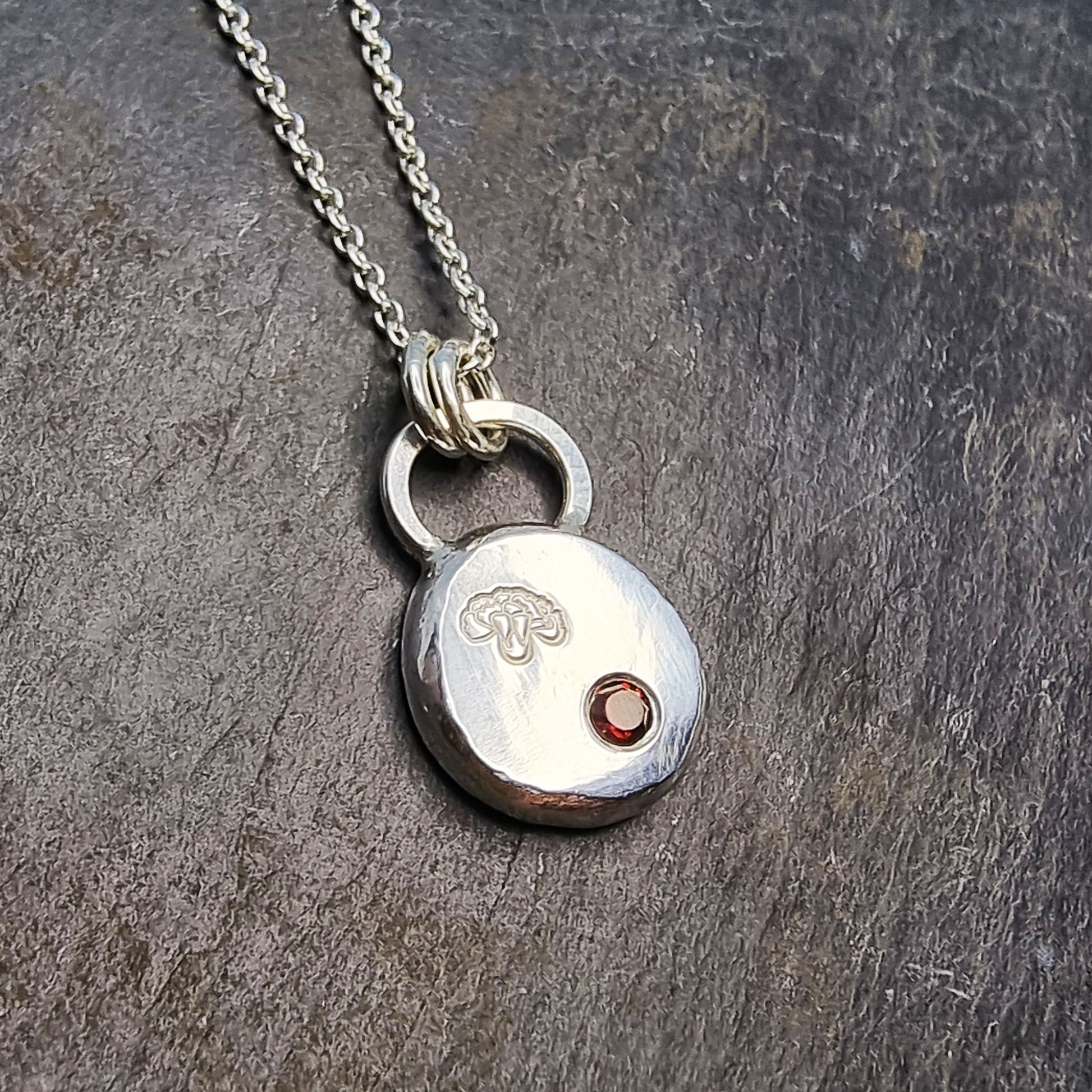 A round silver flat pebble pendant with a carnation flower engraved on it and a red garnet gemstone flush set into it. On a silver chain.