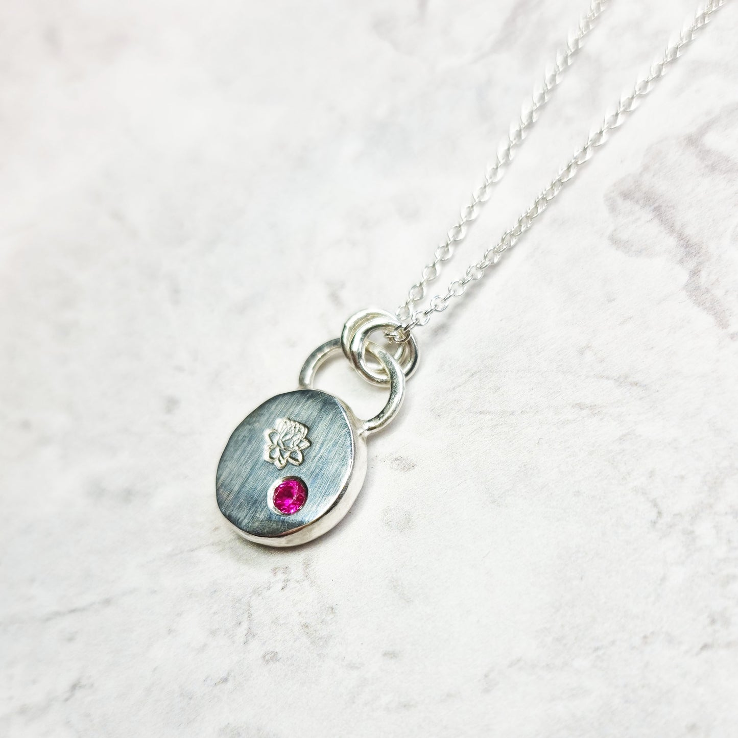 A round silver flat pebble pendant with an engraved lotus flower and flush set red ruby gemstone.