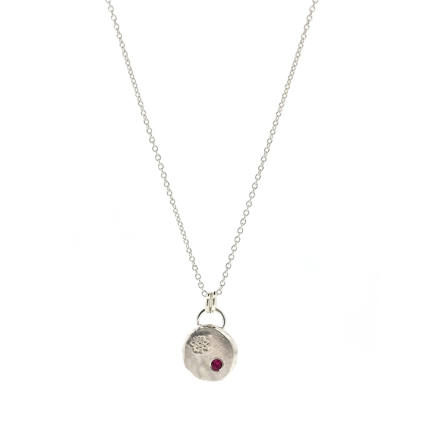 A round silver flat pebble pendant with an engraved lotus flower and flush set red ruby gemstone.