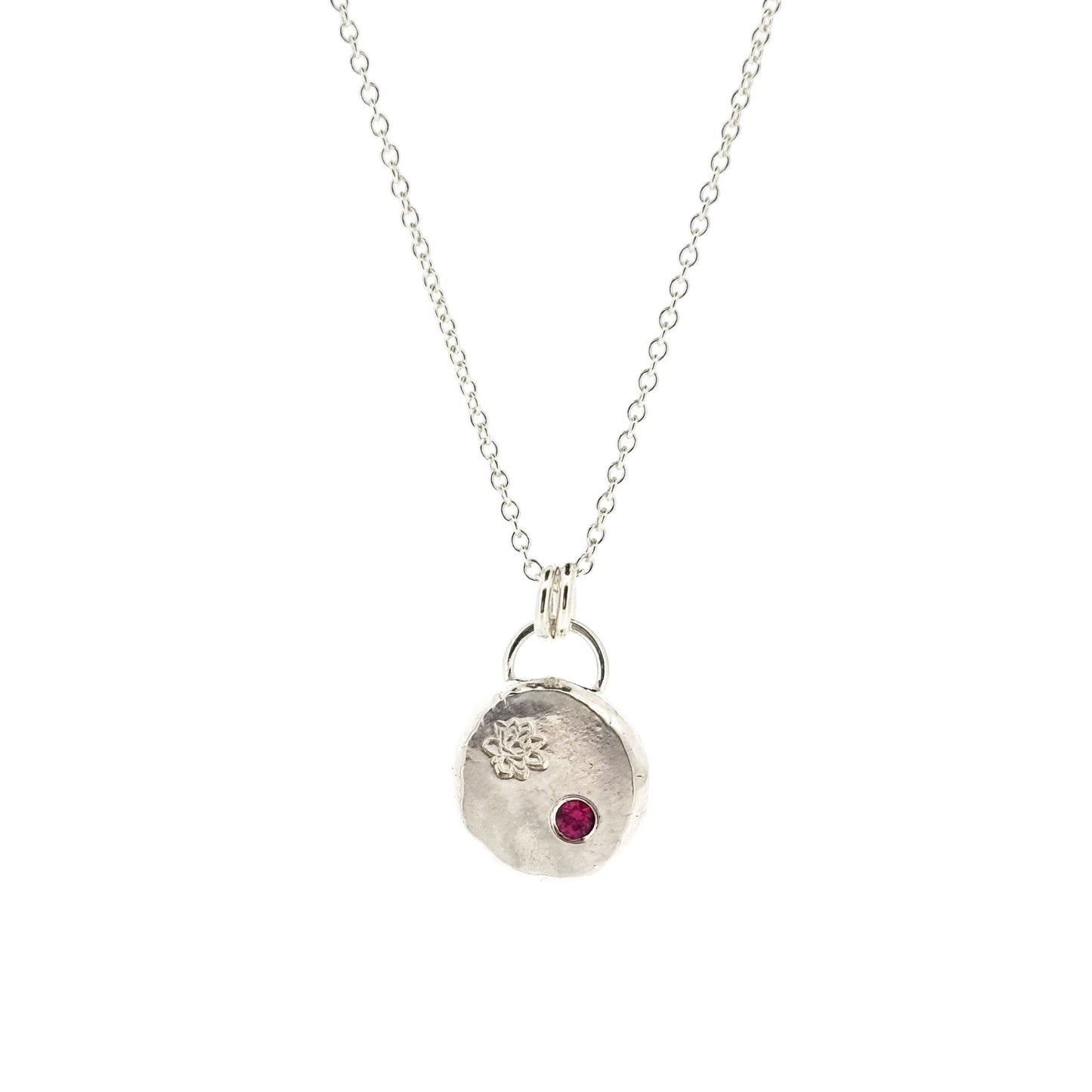 A round silver flat pebble pendant with an engraved lotus flower and flush set red ruby gemstone.