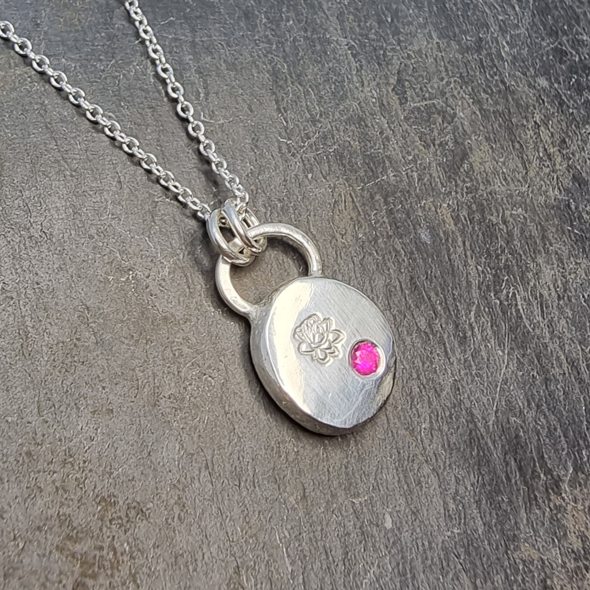 A round silver flat pebble pendant with an engraved lotus flower and flush set red ruby gemstone.