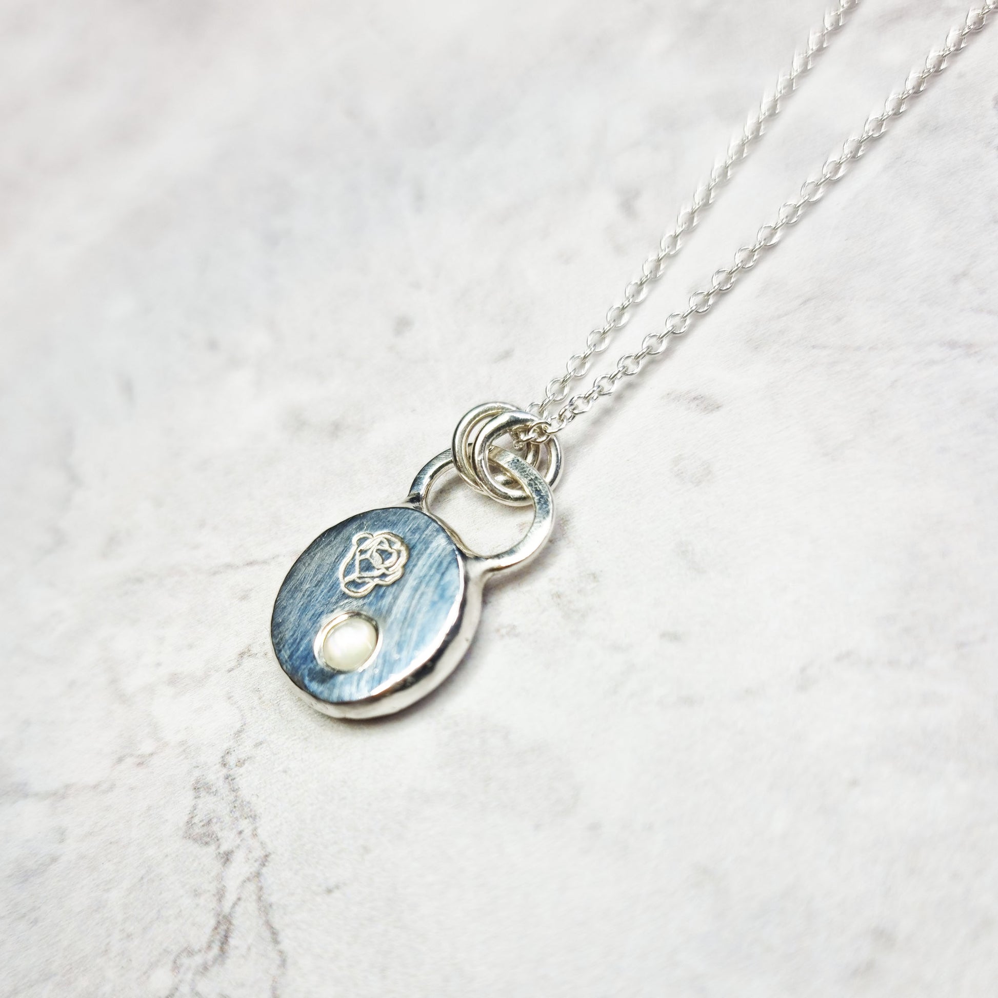 A round silver flat pebble pendant necklace with a rose engraved on it and a flush-set mother of pearl gemstone.