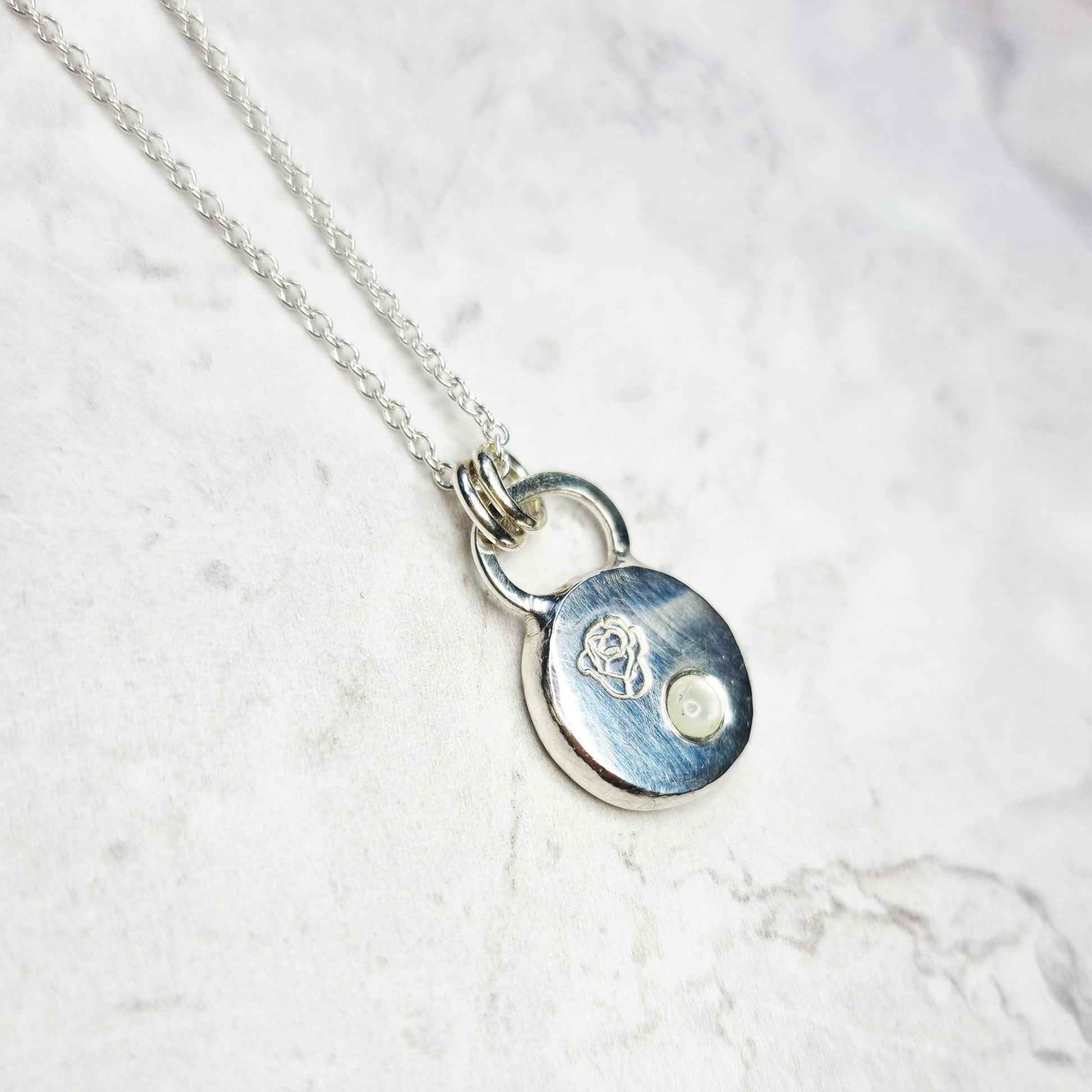 A round silver flat pebble pendant necklace with a rose engraved on it and a flush-set mother of pearl gemstone.