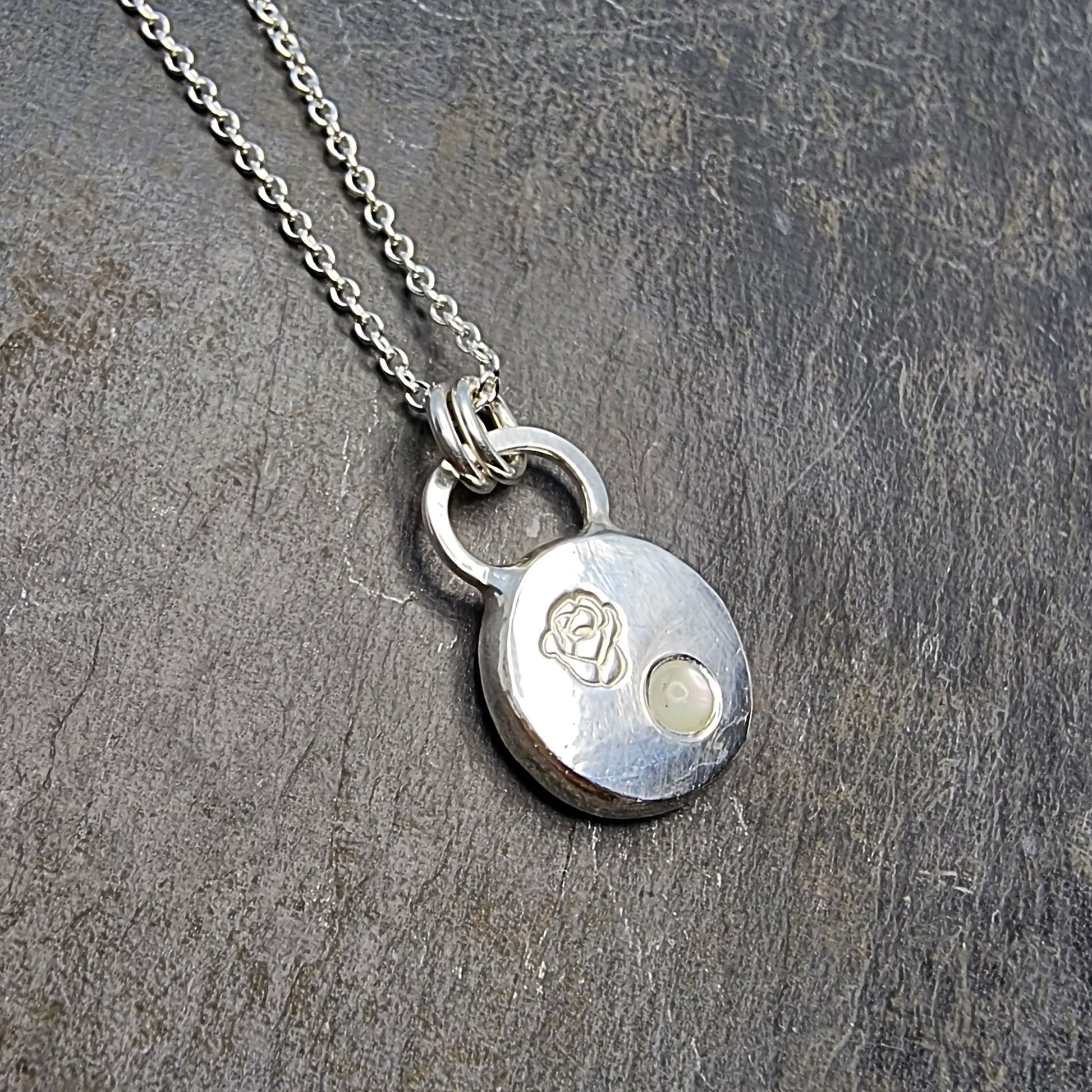 A round silver flat pebble pendant necklace with a rose engraved on it and a flush-set mother of pearl gemstone.