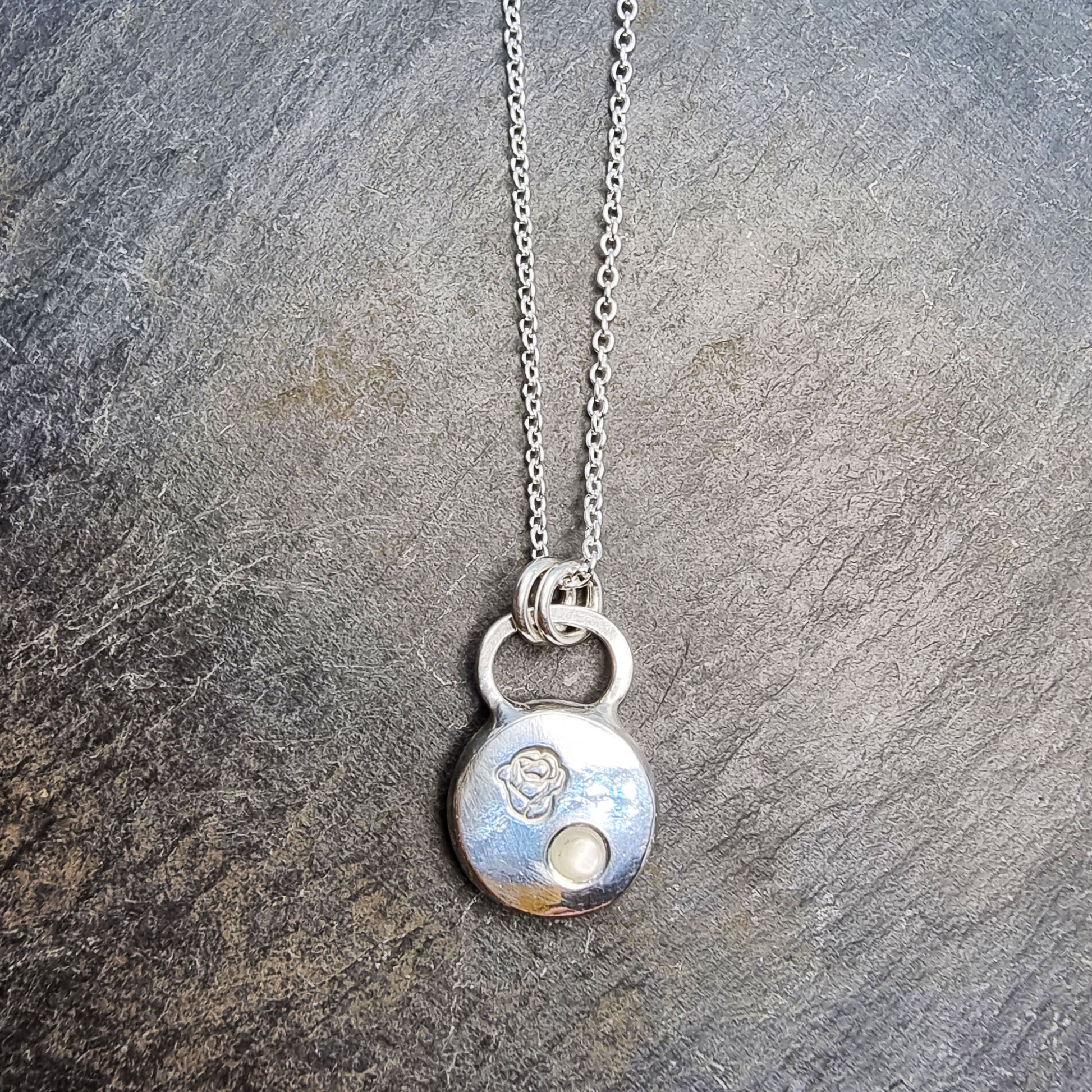 A round silver flat pebble pendant necklace with a rose engraved on it and a flush-set mother of pearl gemstone.