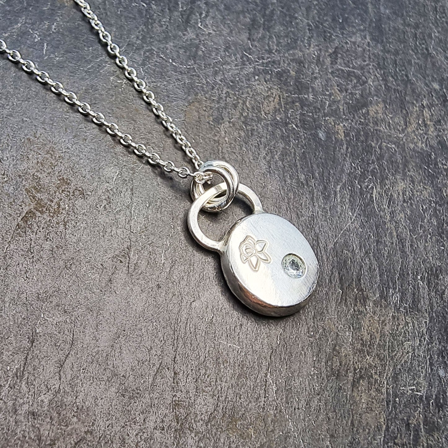 A round silver flat pebble pendant with a daffodil engraved on it and a flush set pale blue aquamarine gemstone.