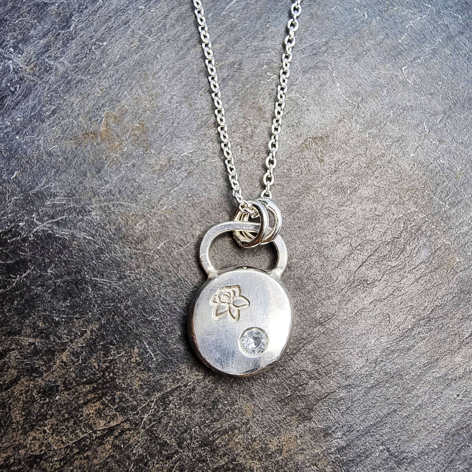 A round silver flat pebble pendant with a daffodil engraved on it and a flush set pale blue aquamarine gemstone.