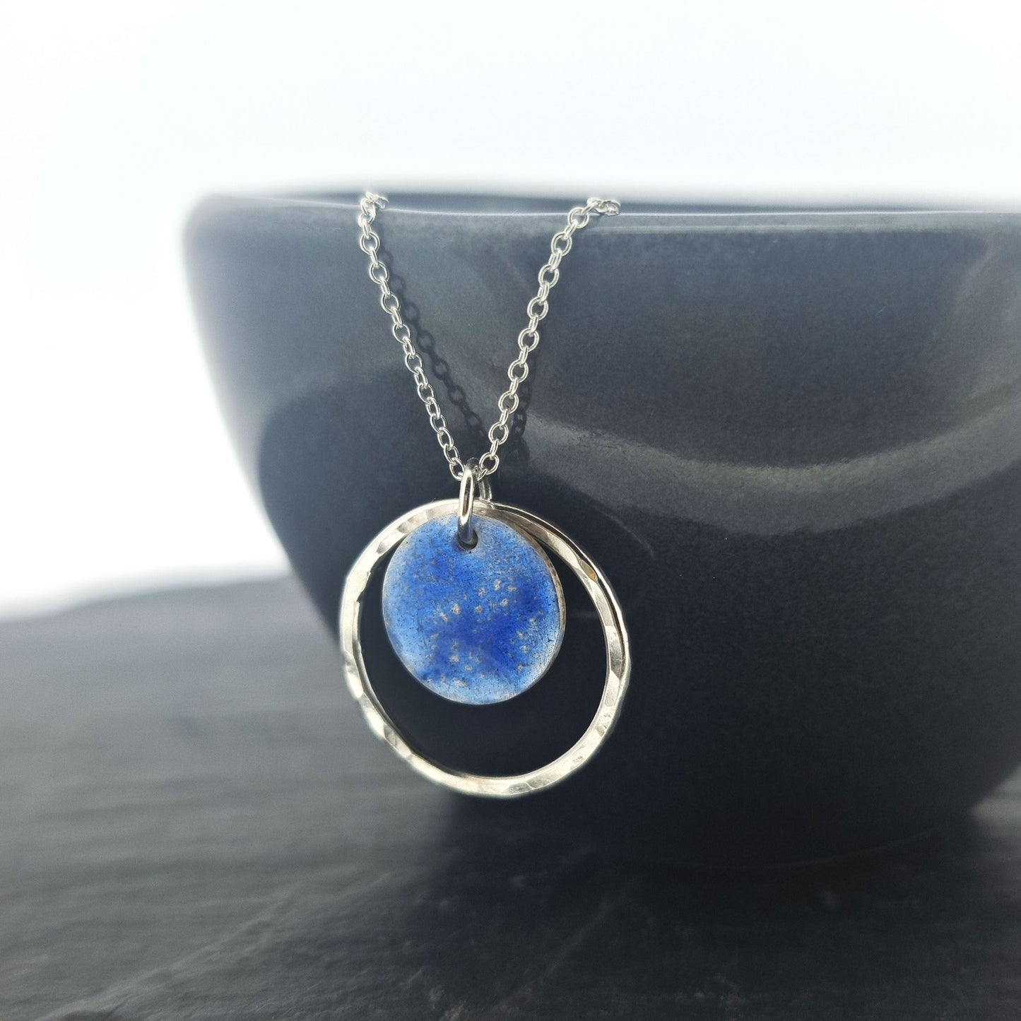 A silver pendant with a central disc with a blue patterned enamel surrounded by a silver hammered open circle. On a silver chain. Pictured on a black bowl.