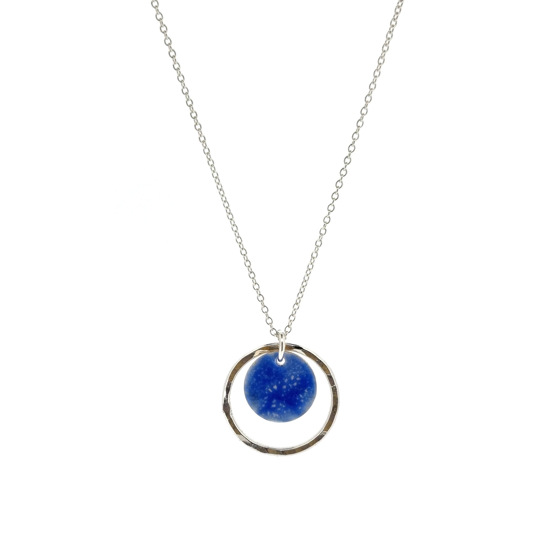 A silver pendant with a central disc with a blue patterned enamel surrounded by a silver hammered open circle. On a silver chain.