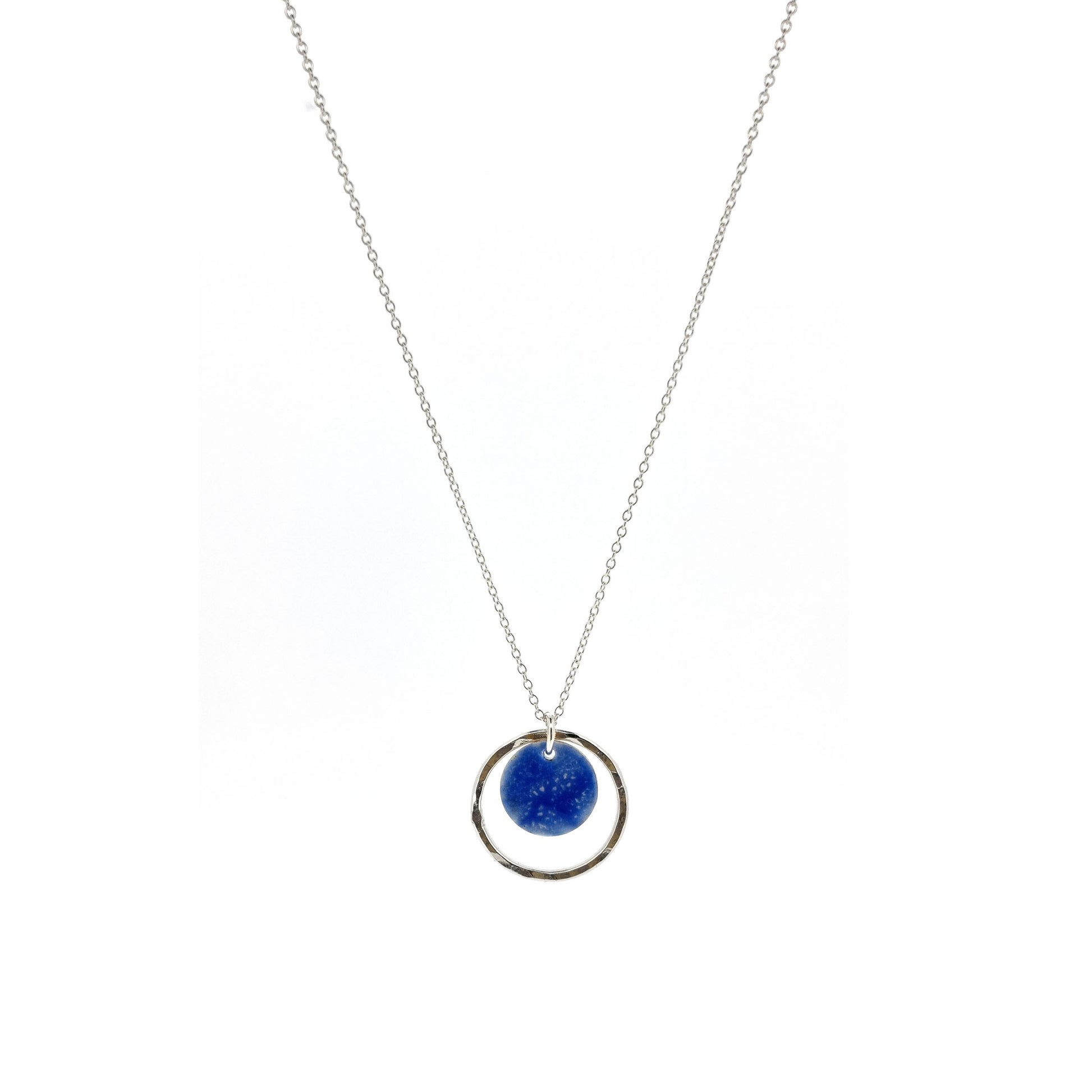 A silver pendant with a central disc with a blue patterned enamel surrounded by a silver hammered open circle. On a silver chain.
