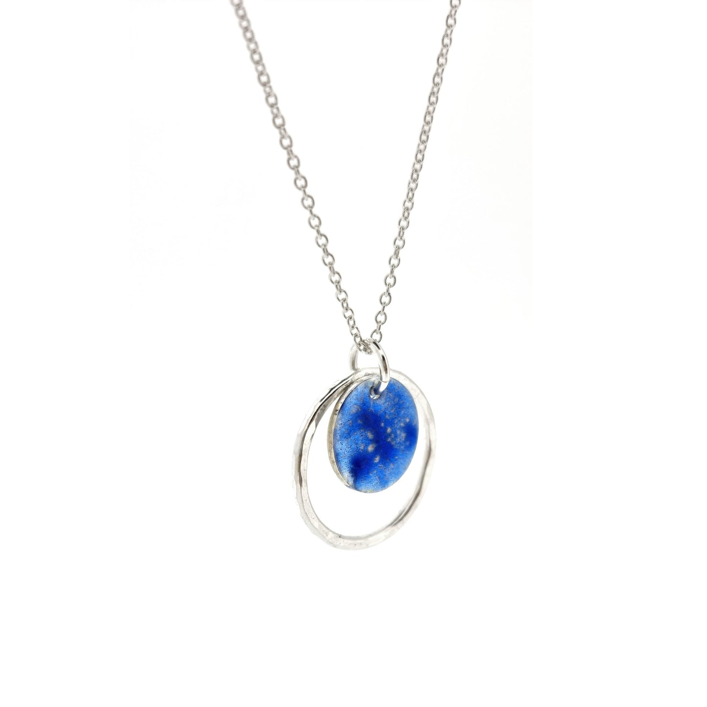 A silver pendant with a central disc with a blue patterned enamel surrounded by a silver hammered open circle. On a silver chain. Side view.