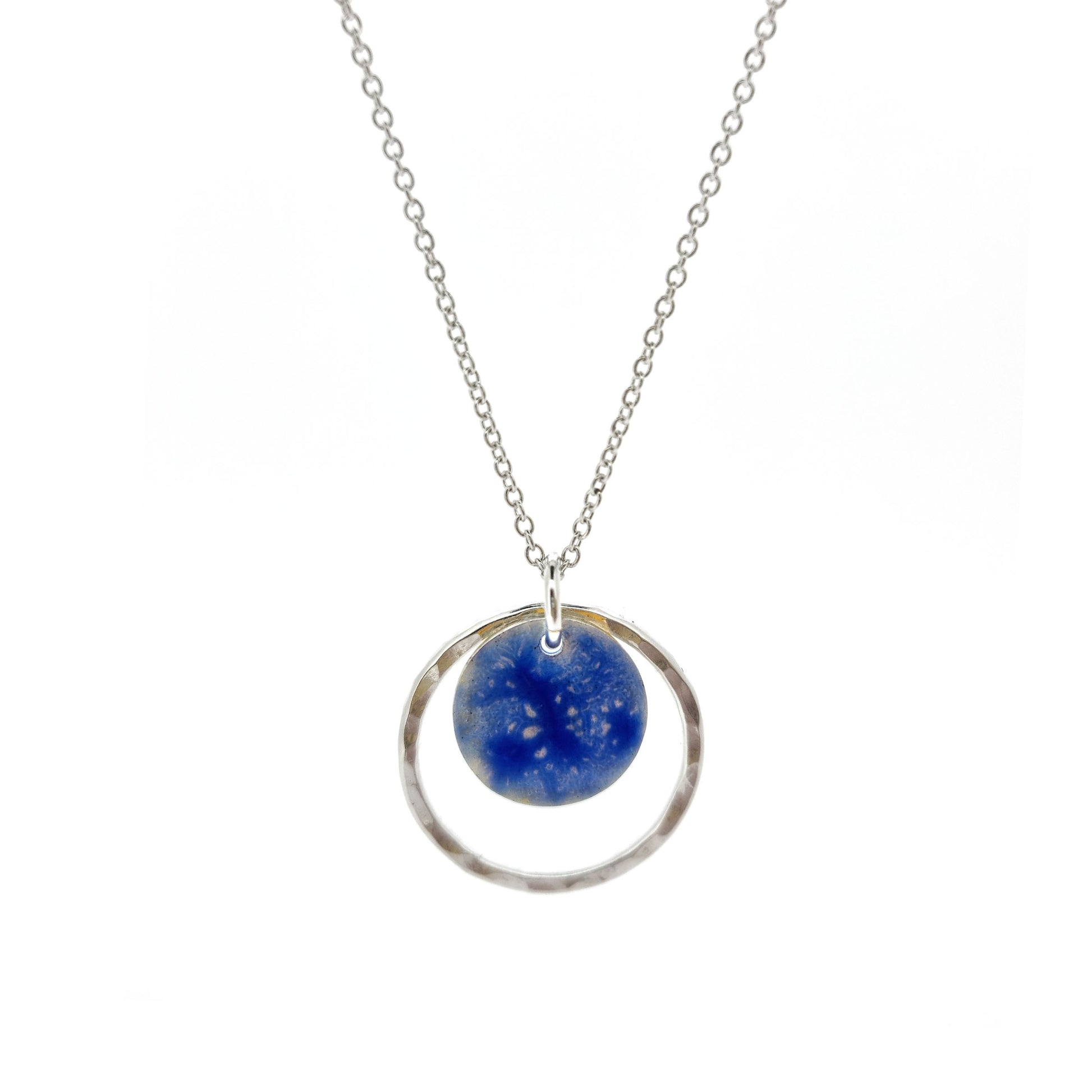 A silver pendant with a central disc with a blue patterned enamel surrounded by a silver hammered open circle. On a silver chain.