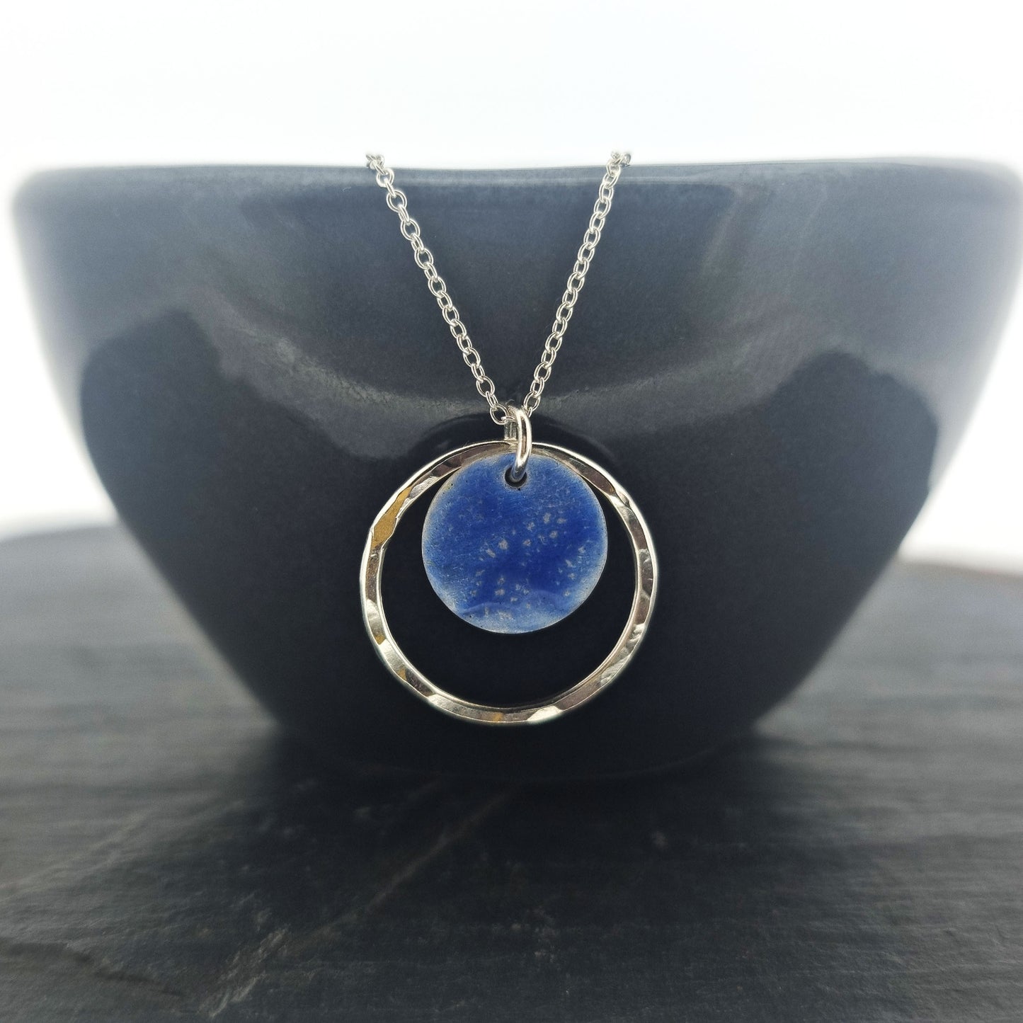 A silver pendant with a central disc with a blue patterned enamel surrounded by a silver hammered open circle. On a silver chain. Pictured on a black bowl.
