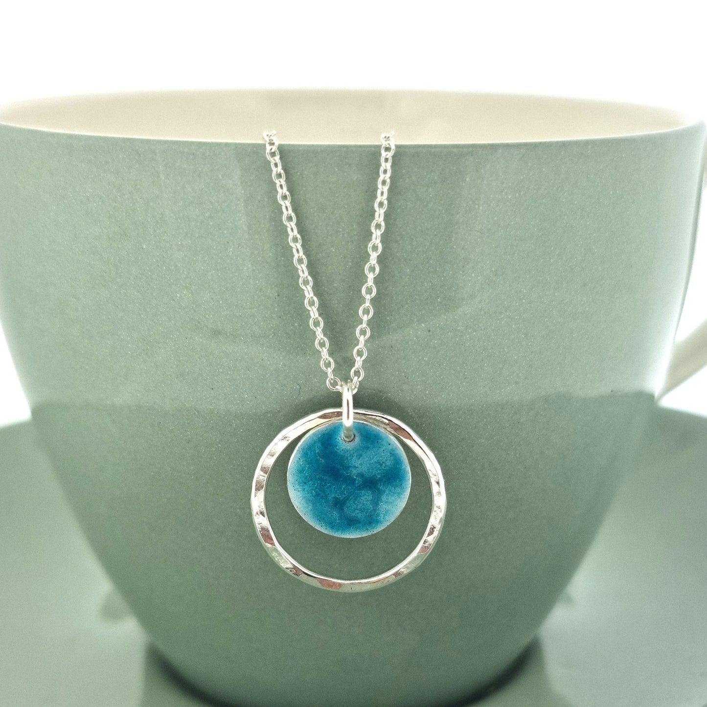 Silver pendant with a central silver disc covered with a turquoise enamel pattern and a surrounding hammered silver circle. On a silver chain. Shown on a cup.