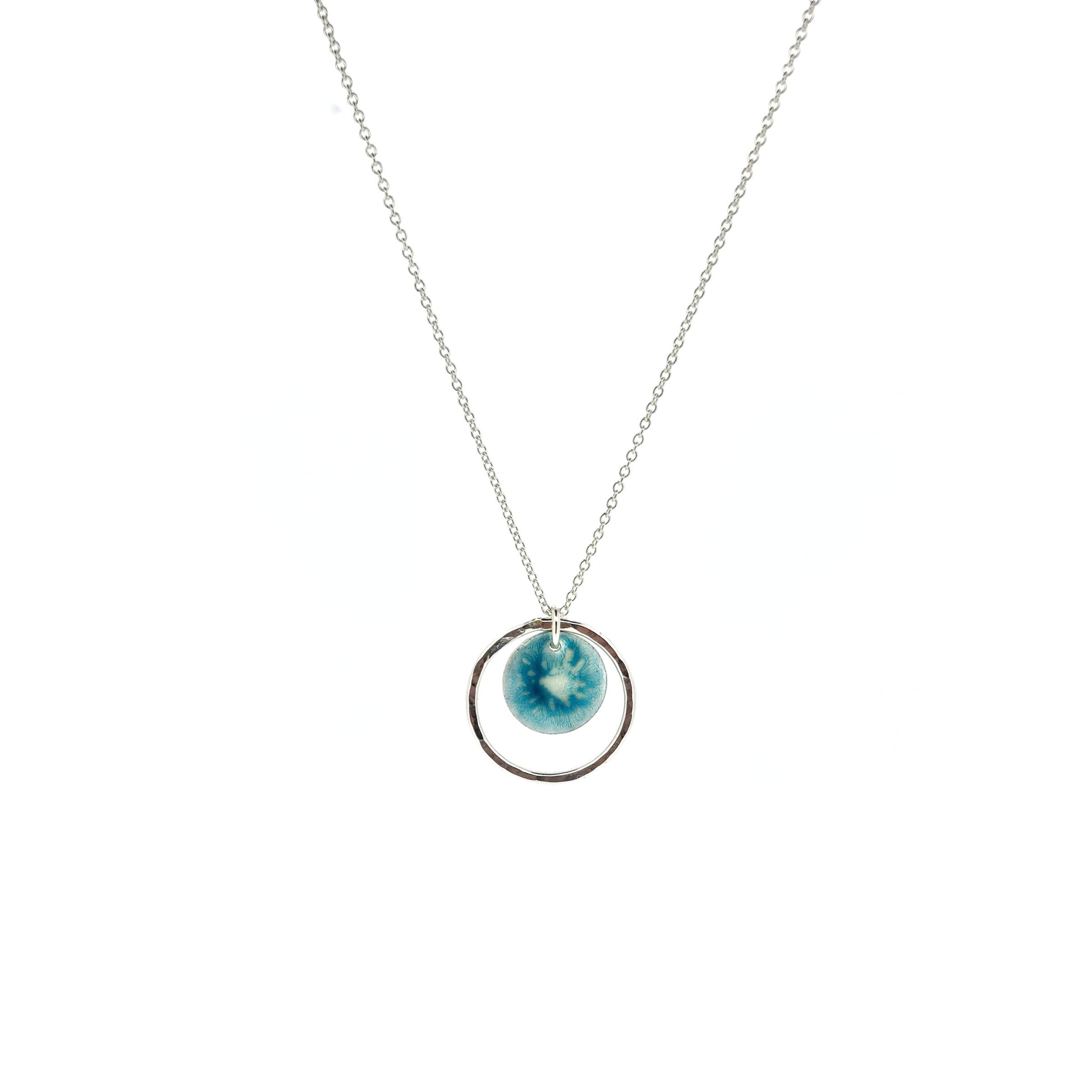 Silver pendant with a central silver disc covered with a turquoise enamel pattern and a surrounding hammered silver circle. On a silver chain.