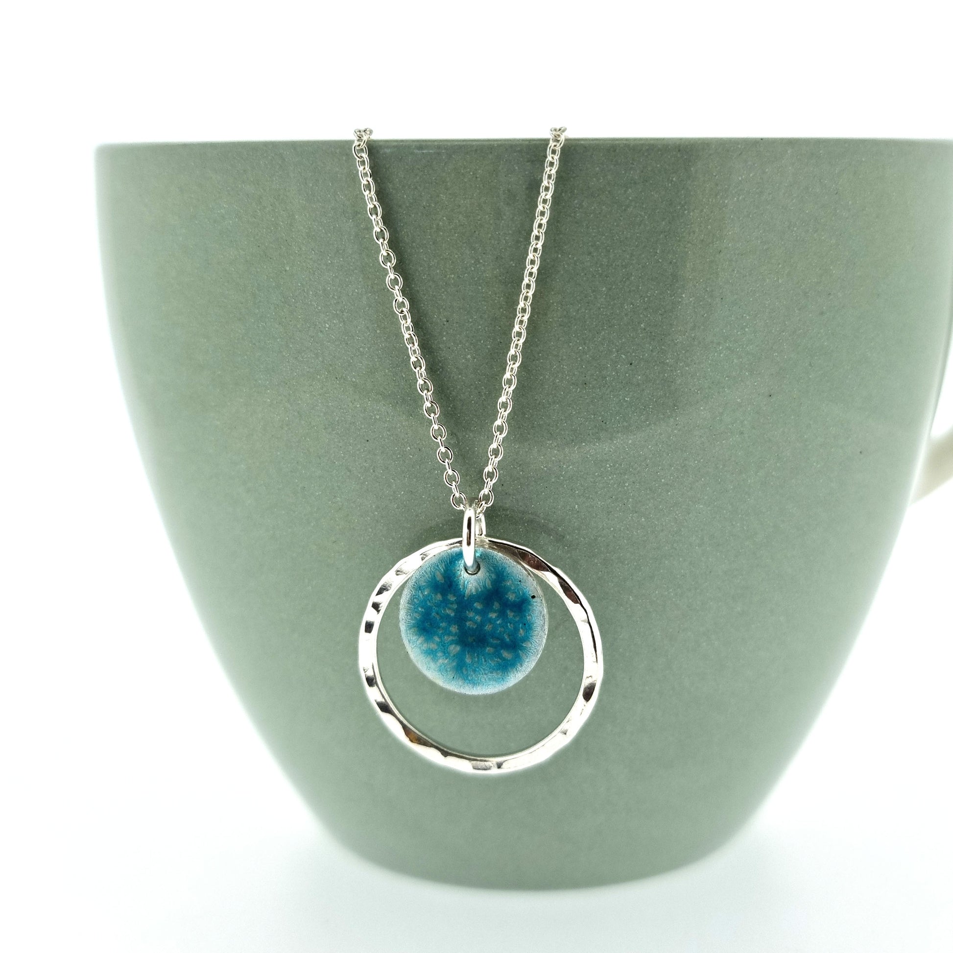 Silver pendant with a central silver disc covered with a turquoise enamel pattern and a surrounding hammered silver circle. On a silver chain. Pictured on cup.