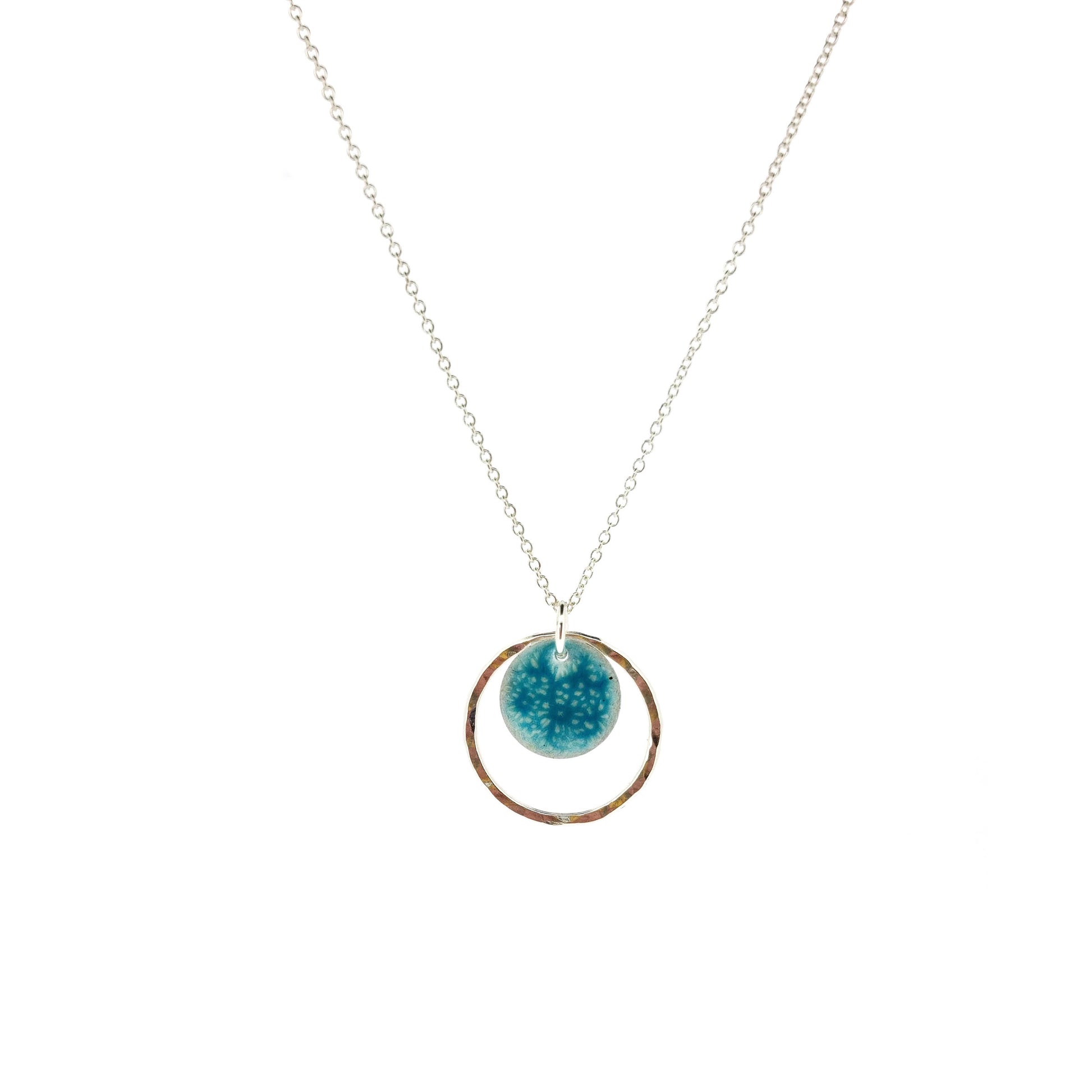 Silver pendant with a central silver disc covered with a turquoise enamel pattern and a surrounding hammered silver circle. On a silver chain.