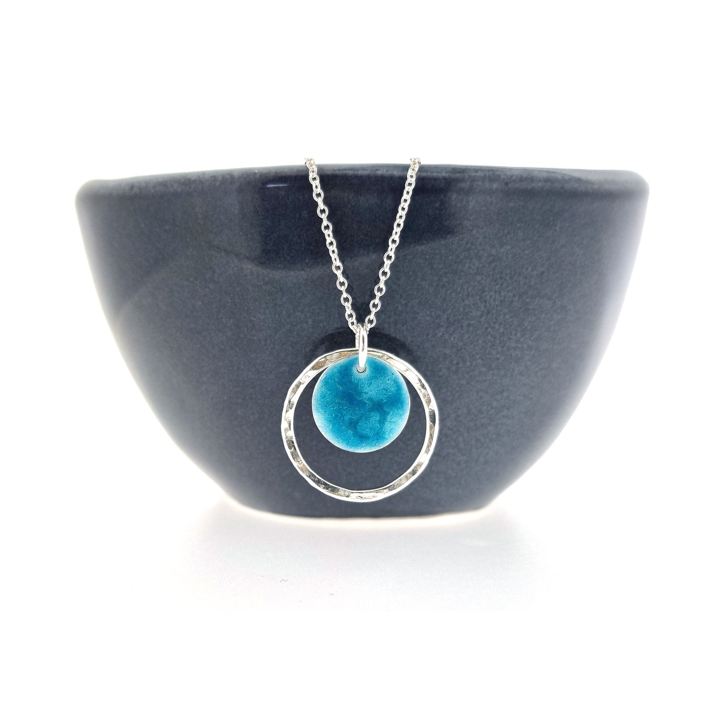 Silver pendant with a central silver disc covered with a turquoise enamel pattern and a surrounding hammered silver circle. On a silver chain. Shown on a bowl.