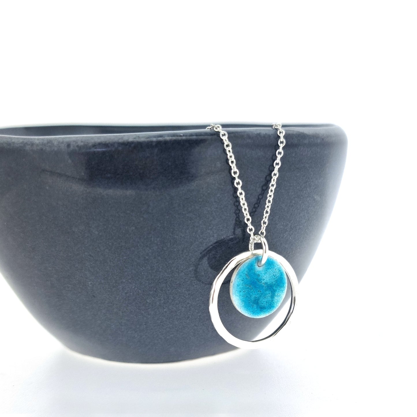 Silver pendant with a central silver disc covered with a turquoise enamel pattern and a surrounding hammered silver circle. On a silver chain. Shown on a bowl.