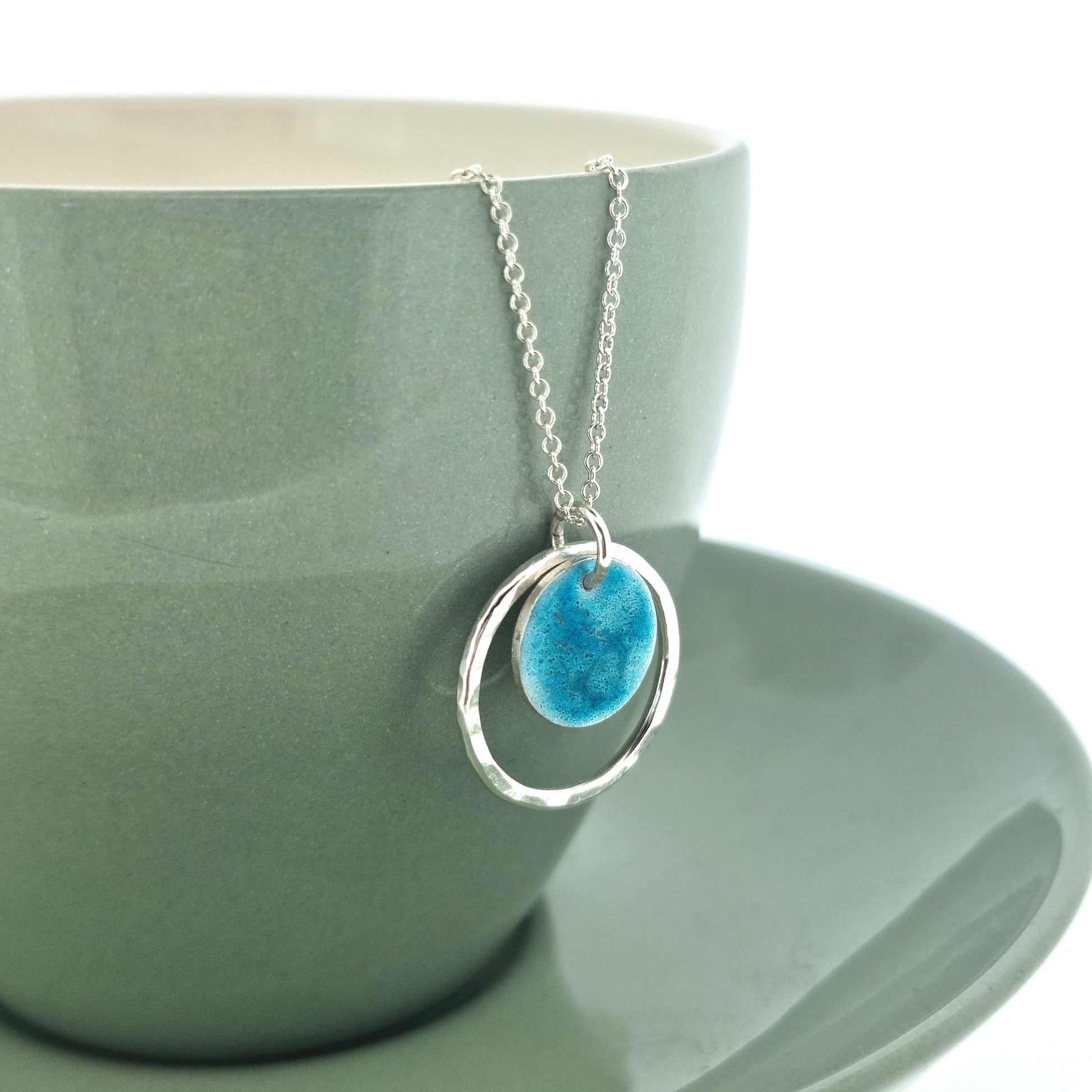 Silver pendant with a central silver disc covered with a turquoise enamel pattern and a surrounding hammered silver circle. On a silver chain. Shown on a cup.