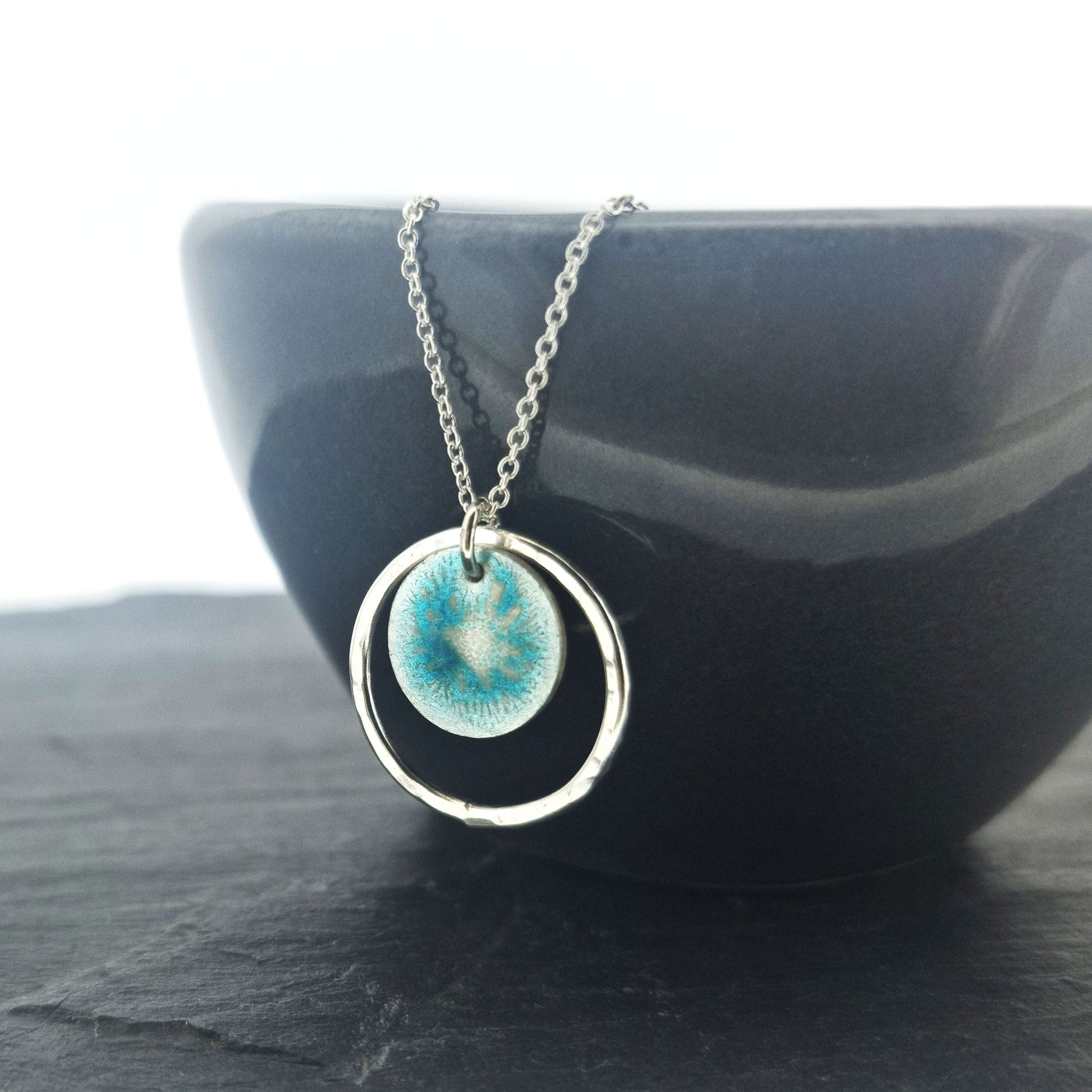 Silver pendant with a central silver disc covered with a turquoise enamel pattern and a surrounding hammered silver circle. On a silver chain. Pictured on a black bowl.