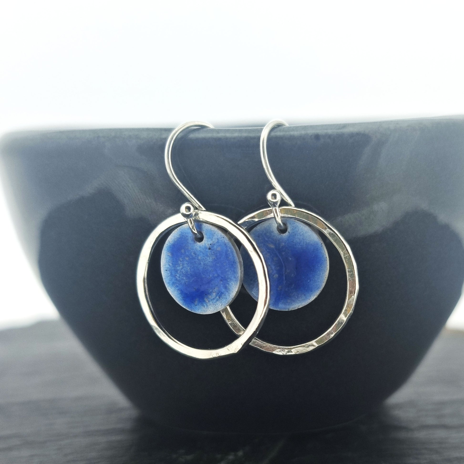 Silver drop earrings with a central disc featuring patterned dark blue enamel surrounded by a silver hammered open circle. Pictured on a dark bowl.