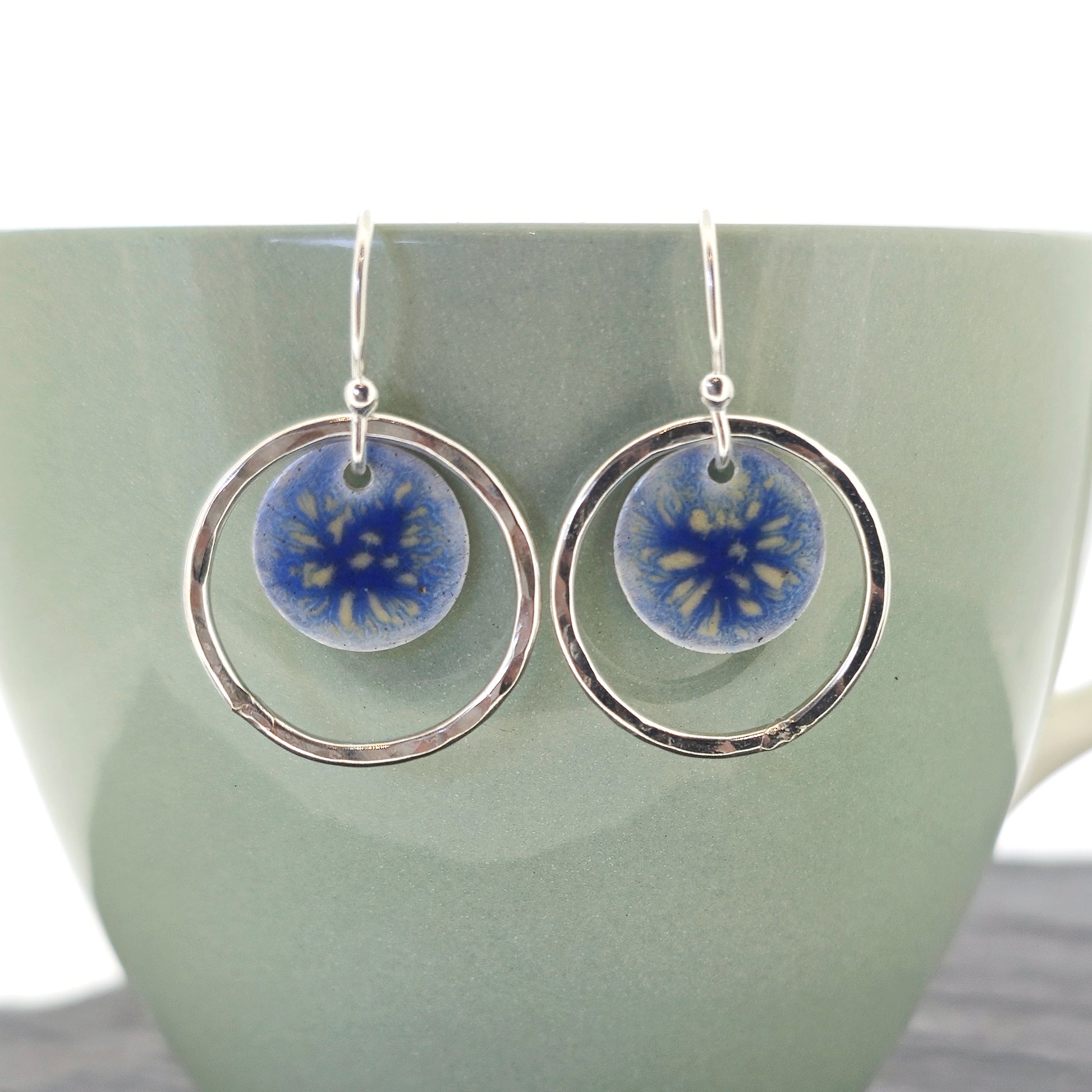 Silver drop earrings with a central disc featuring patterned dark blue enamel surrounded by a silver hammered open circle.  Shown on a cup.