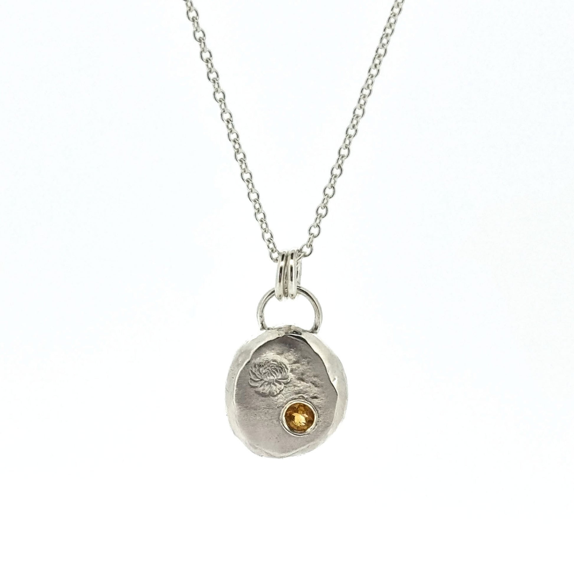 Round silver flat pebble pendant with chrysanthemum flower engraved on it and a flush set yellow citrine gemstone. On a silver chain.