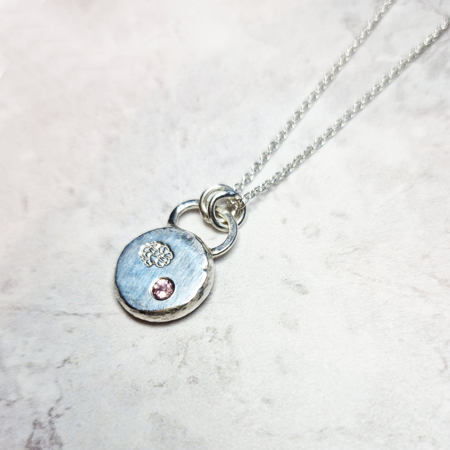 Round silver pendant with an engraved marigold flower and a flush set pink tourmaline gemstone. On a silver chain.