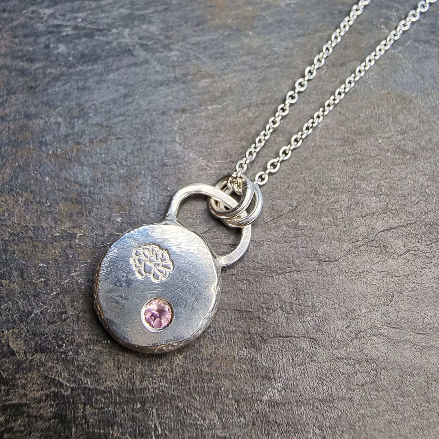 Round silver pendant with an engraved marigold flower and a flush set pink tourmaline gemstone. On a silver chain.