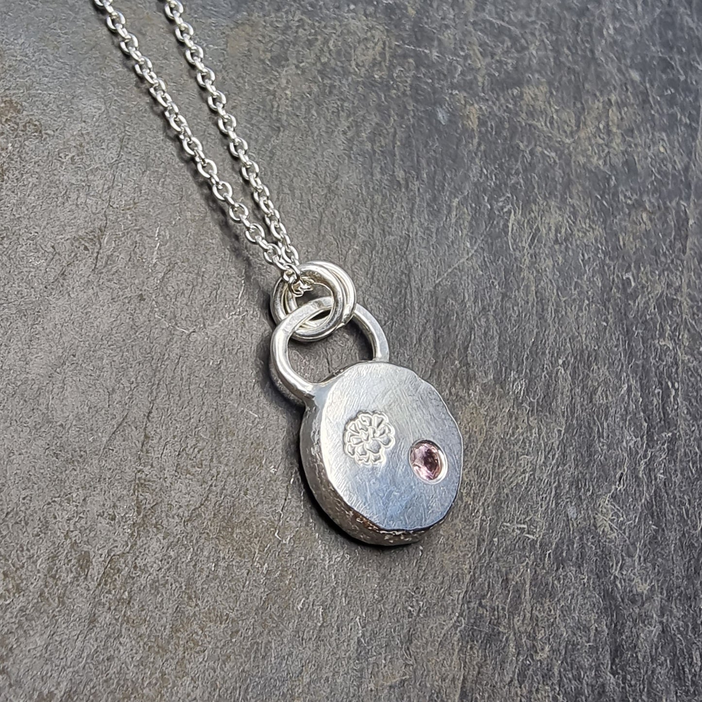 Round silver pendant with an engraved marigold flower and a flush set pink tourmaline gemstone. On a silver chain.