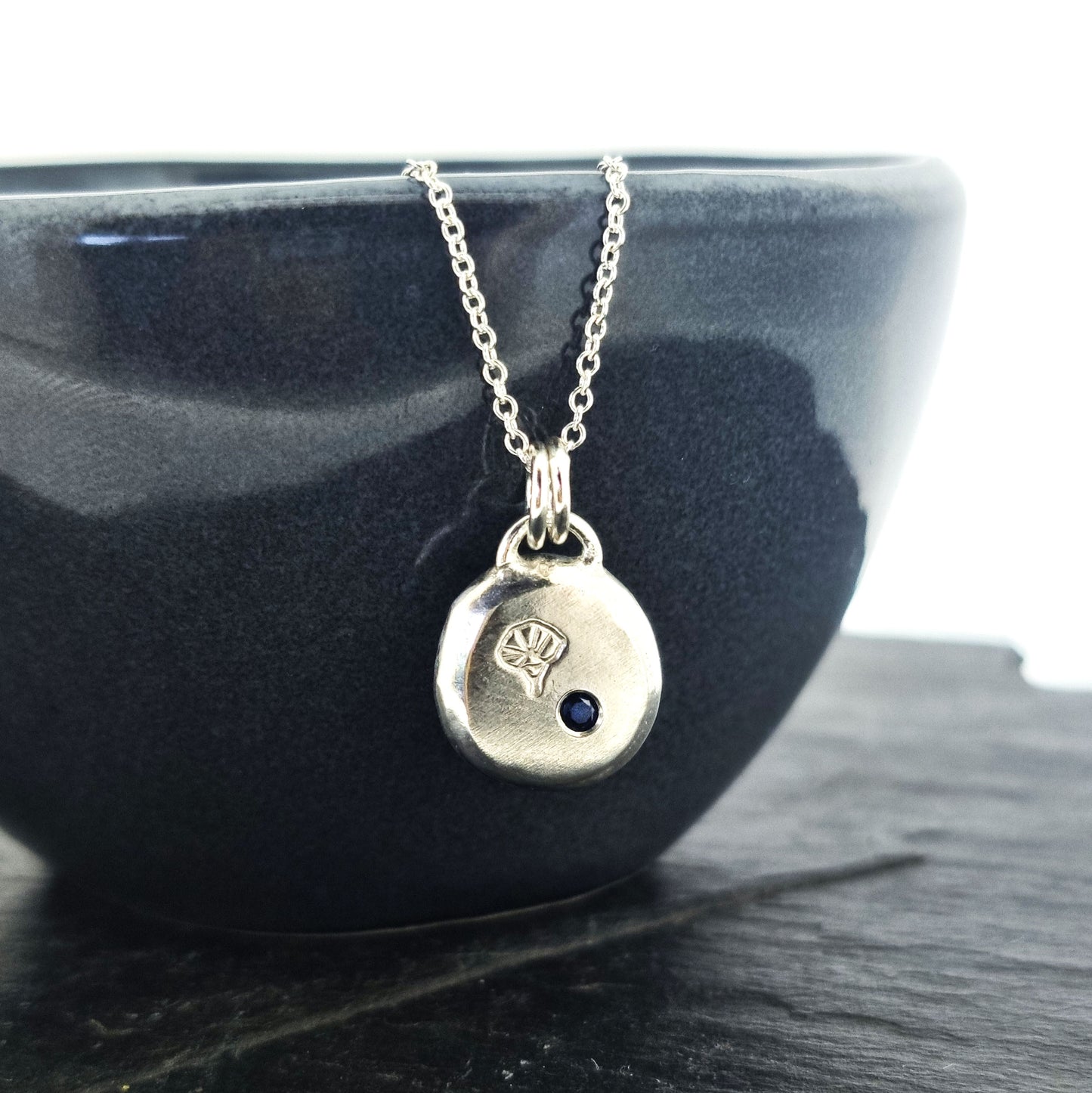 A round flat pebble pendant with a morning glory flower engraved on it and a flush set dark blue sapphire. On a silver chain.