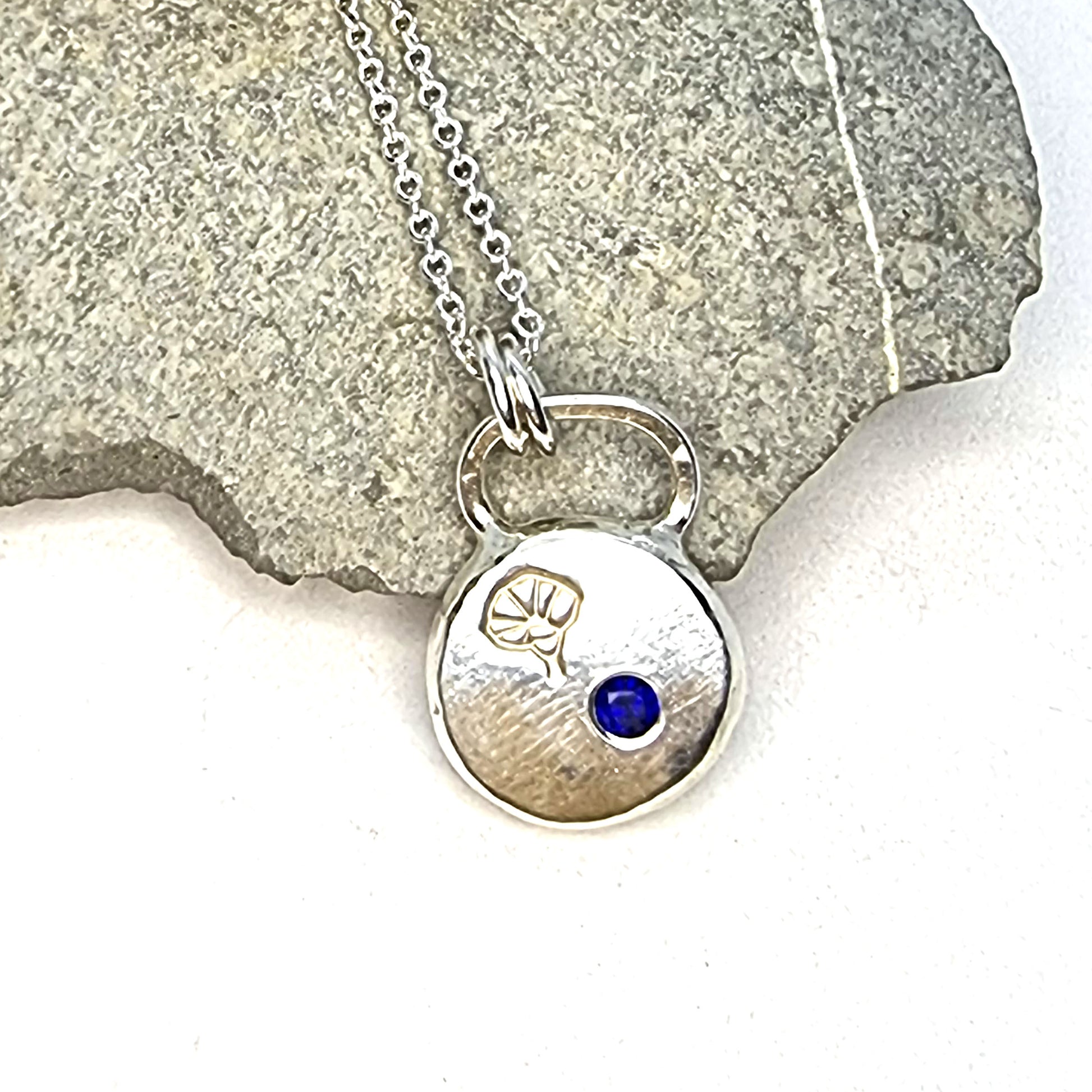 A round flat pebble pendant with a morning glory flower engraved on it and a flush set dark blue sapphire. On a silver chain. Pictured on a stone.