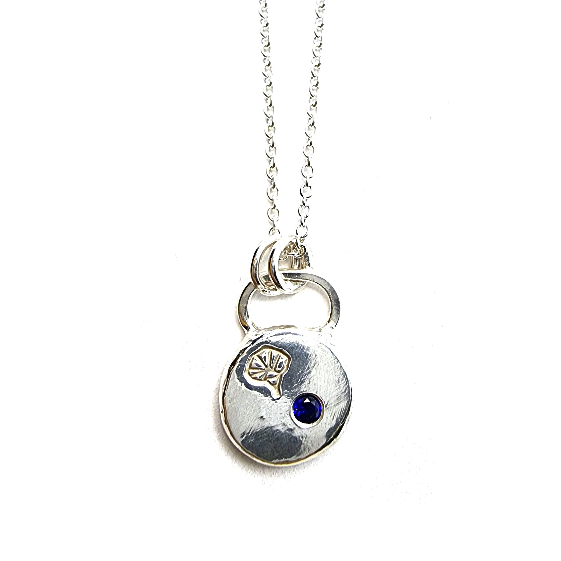 A round flat pebble pendant with a morning glory flower engraved on it and a flush set dark blue sapphire. On a silver chain.