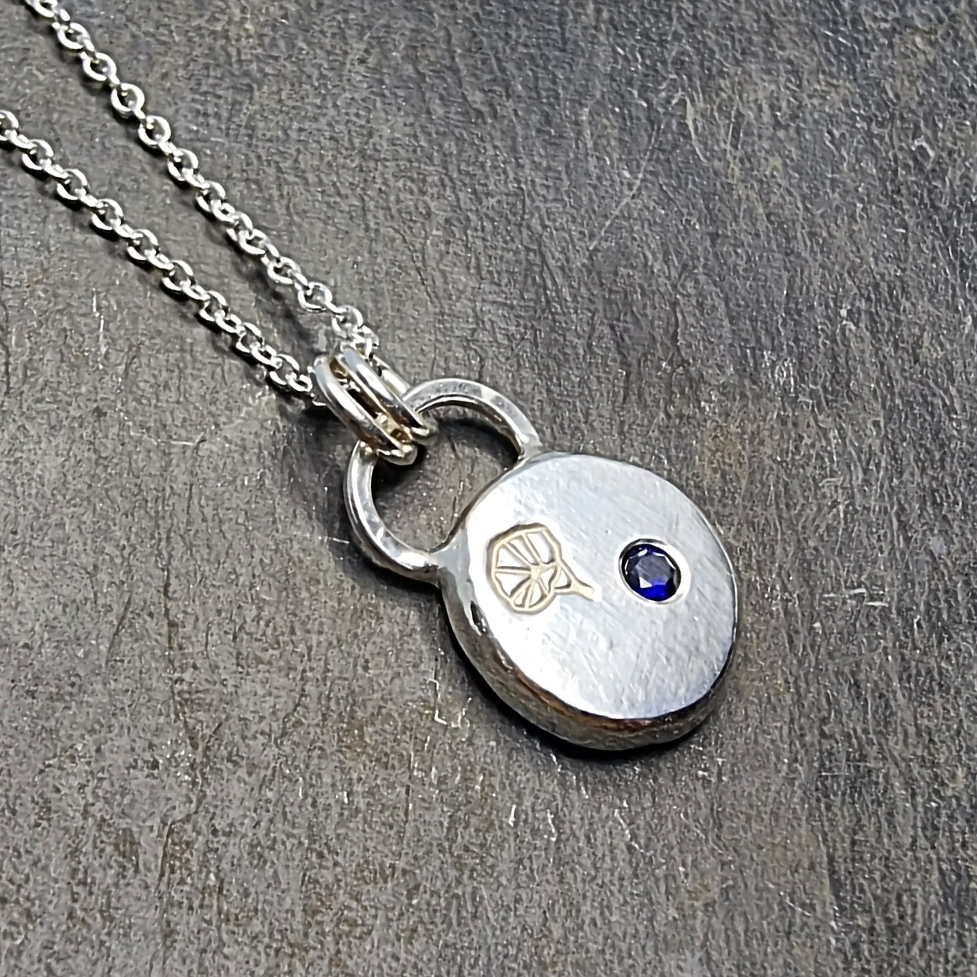 A round flat pebble pendant with a morning glory flower engraved on it and a flush set dark blue sapphire. On a silver chain.