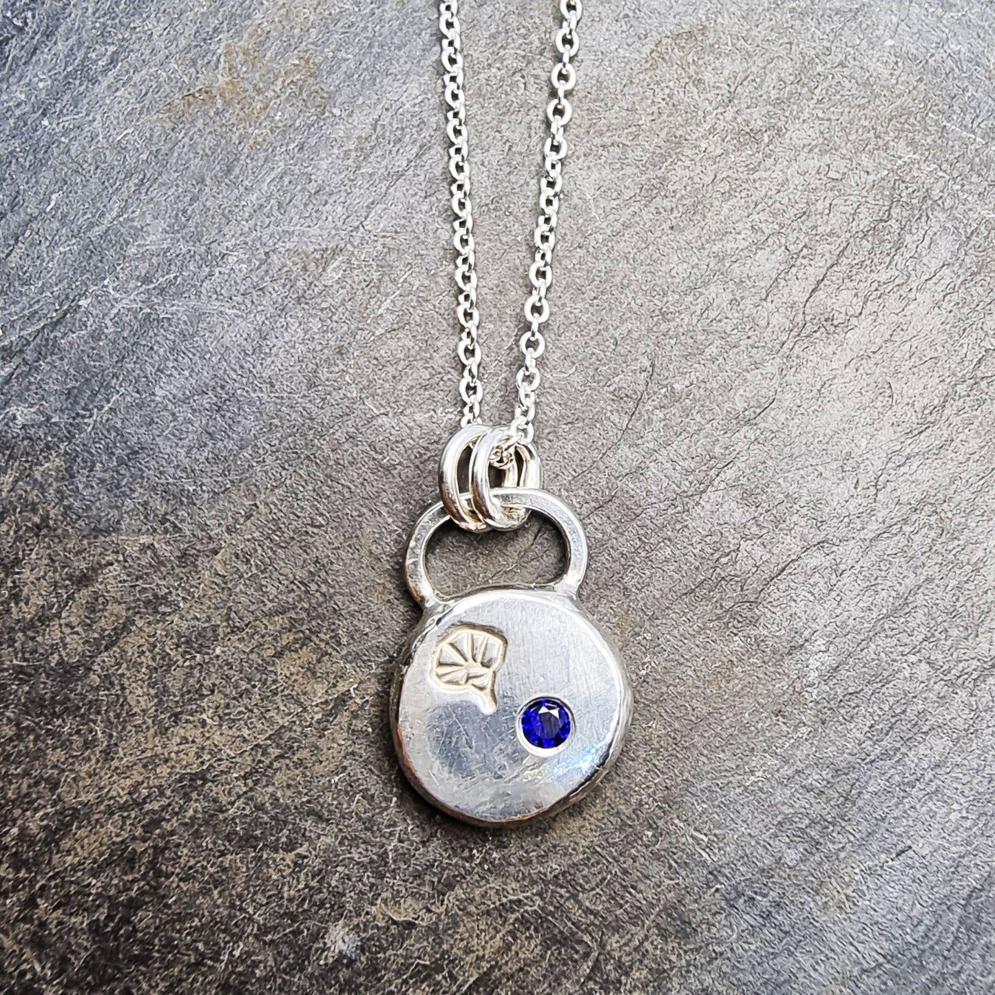 A round flat pebble pendant with a morning glory flower engraved on it and a flush set dark blue sapphire. On a silver chain.