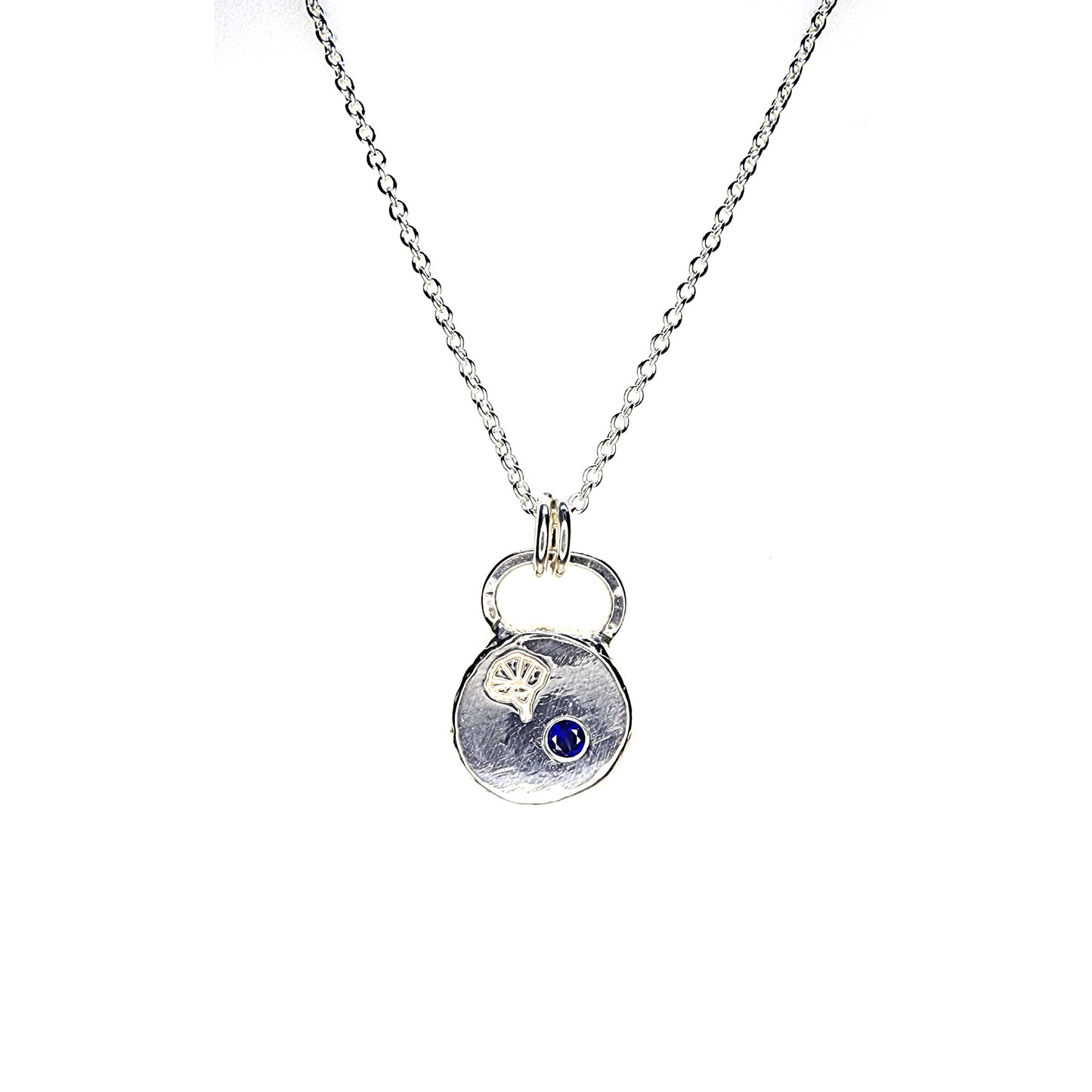 A round flat pebble pendant with a morning glory flower engraved on it and a flush set dark blue sapphire. On a silver chain.