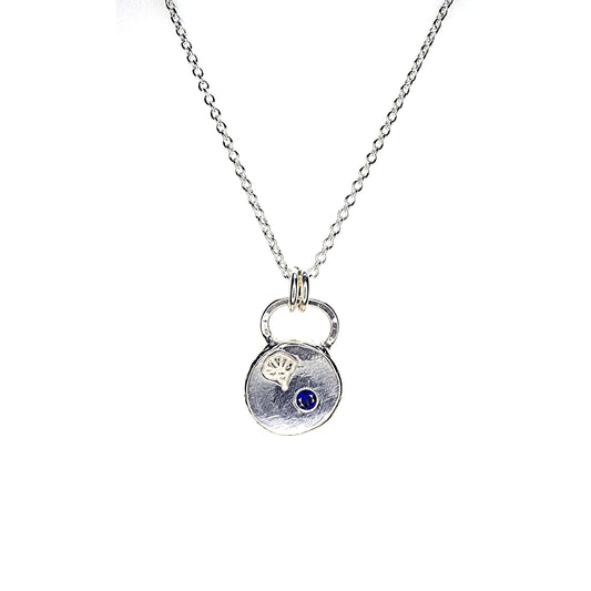 A round flat pebble pendant with a morning glory flower engraved on it and a flush set dark blue sapphire. On a silver chain.